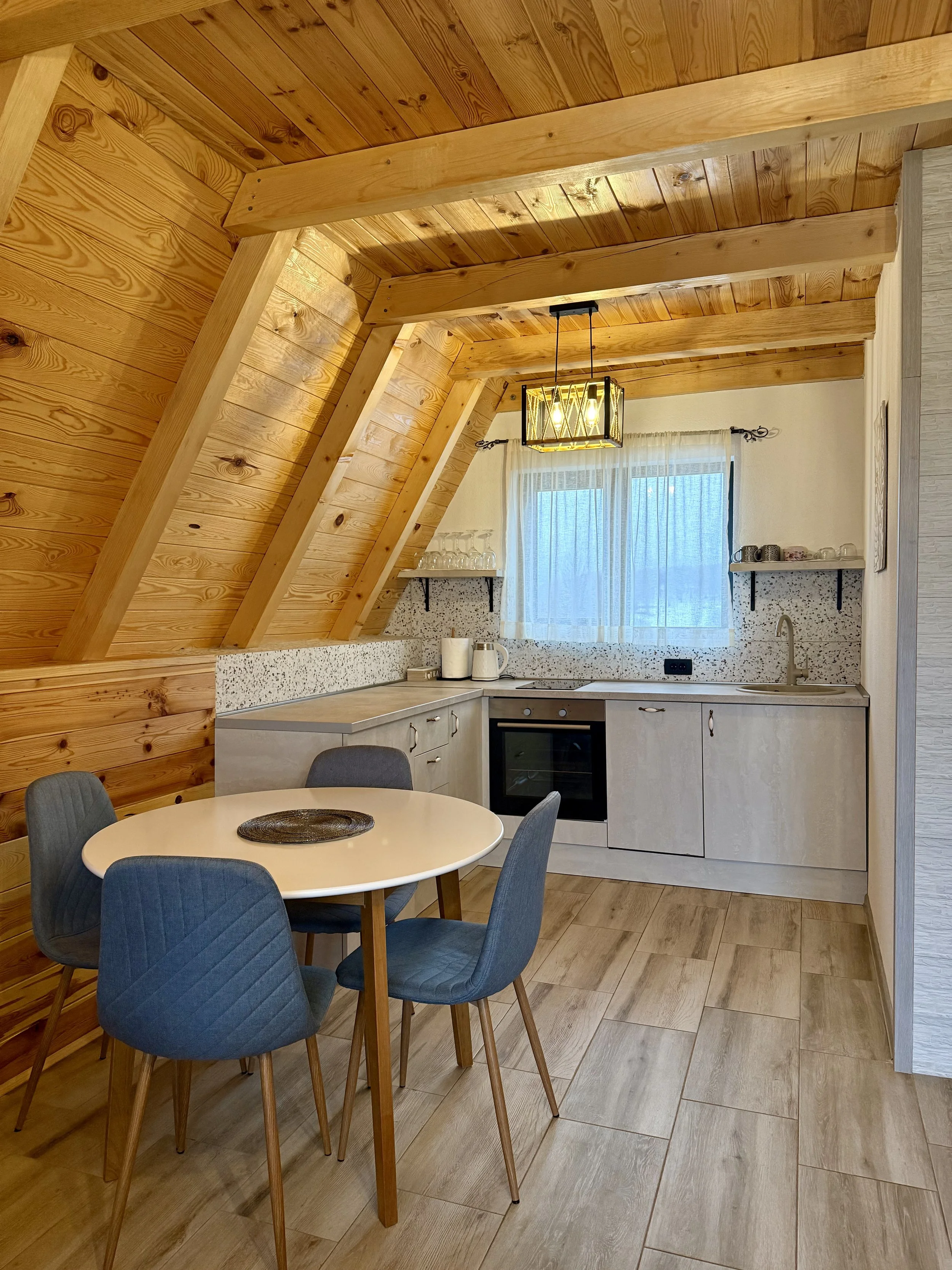A cozy kitchen with wood-paneled slanted ceiling, a round dining table with four blue upholstered chairs, a window with white sheer curtains, and modern light fixtures.