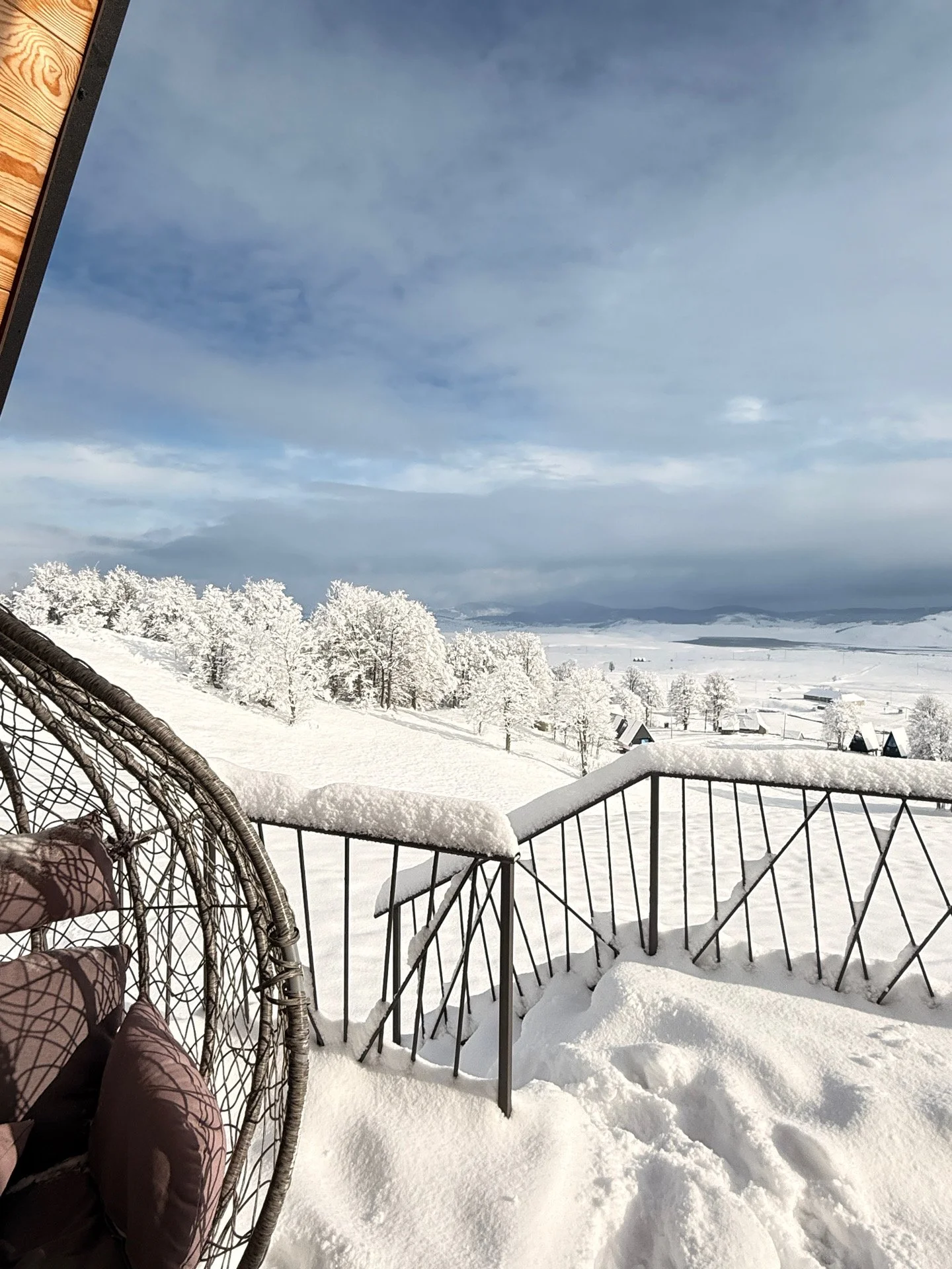 Snow-covered balcony with black metal railing overlooking a winter landscape of snow-covered trees and distant hills under a partly cloudy sky.