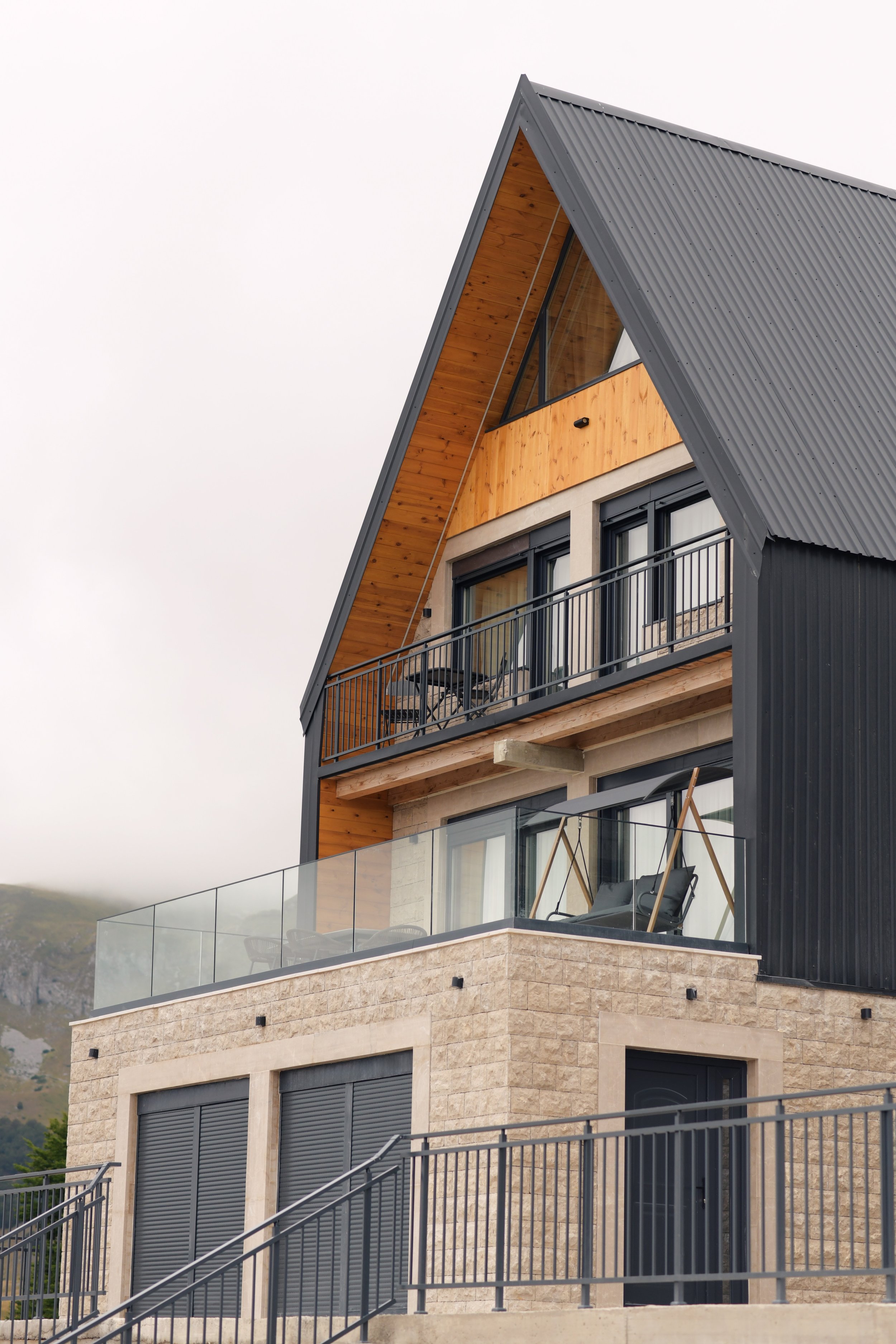 Modern multi-story house with a beige brick base, black railing balconies, and dark metal roofing, surrounded by mountains.