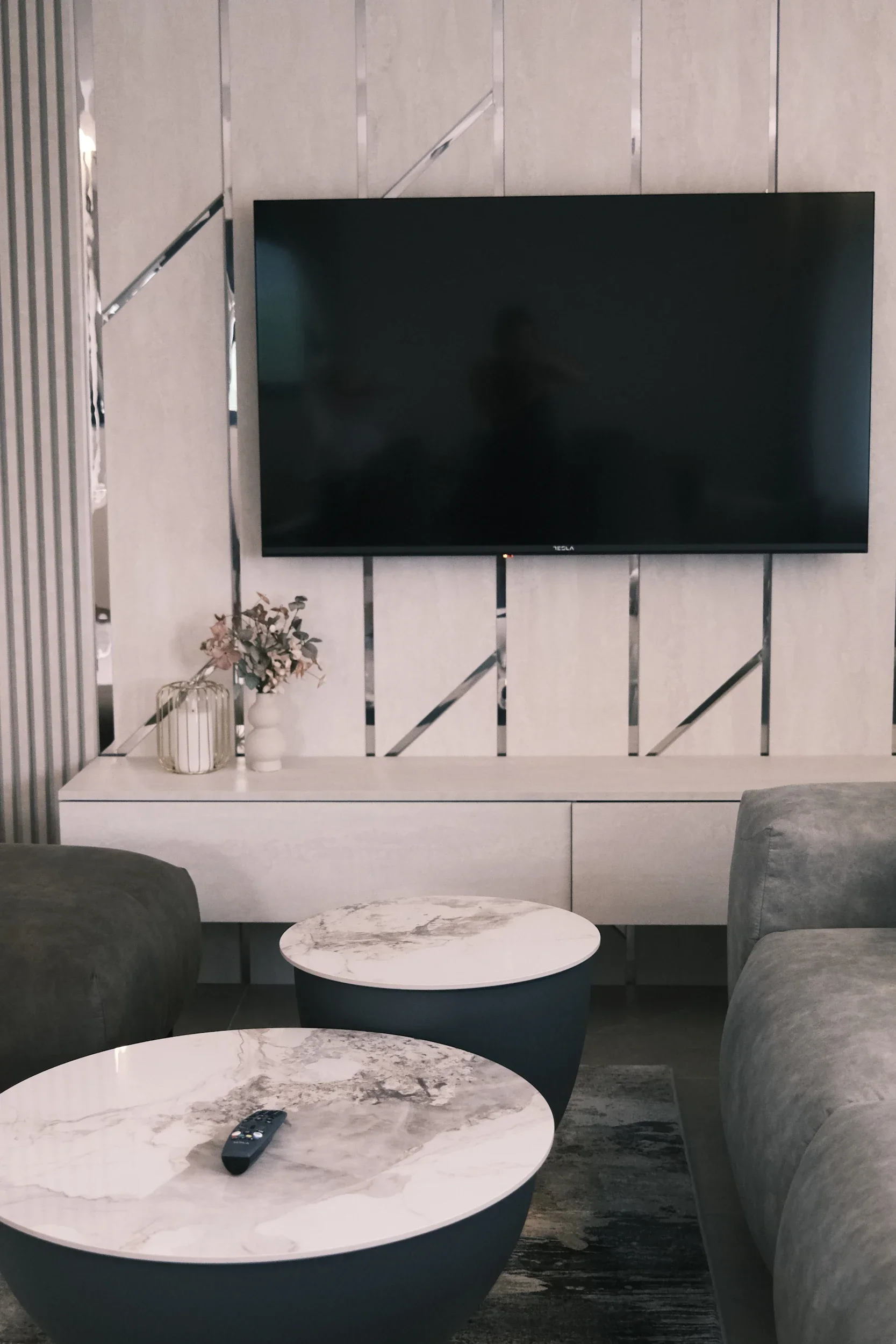 Living room with a mounted flat-screen TV, a white cabinet below, two marble-topped coffee tables, gray couches, and decorative items on the cabinet including a vase with flowers and a small lantern.