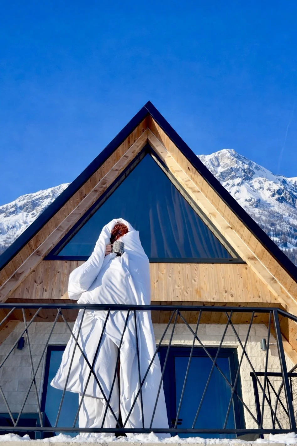 A person wrapped in a white blanket on a balcony of a house with wood siding, holding a mug, with snow-covered mountains in the background under a clear blue sky.