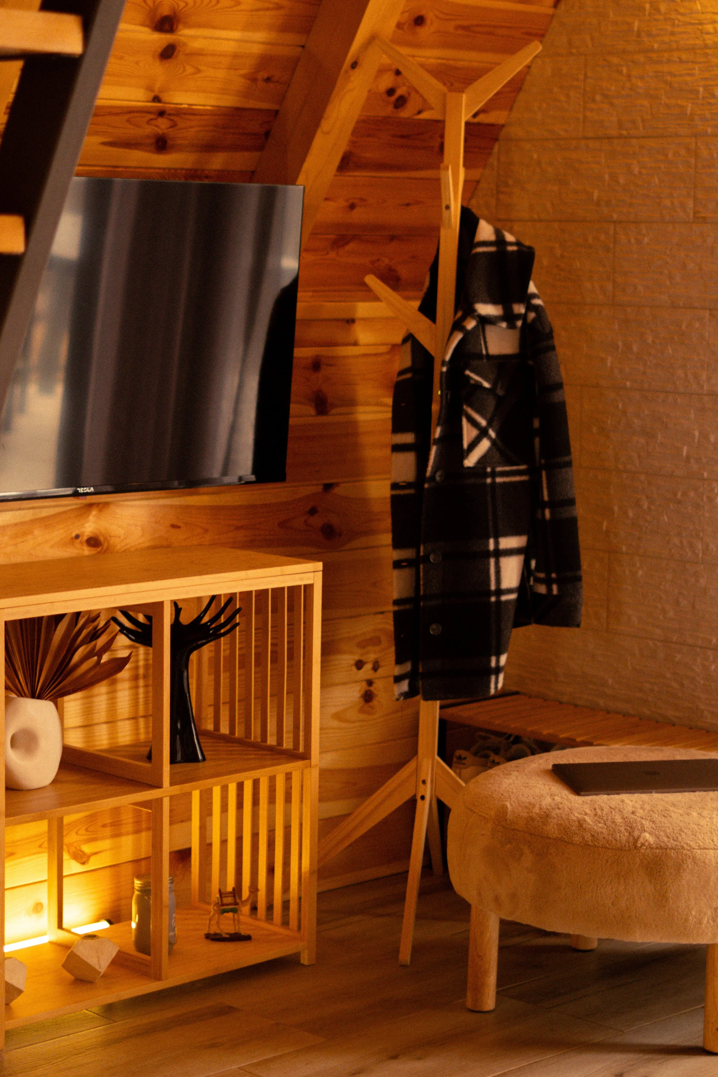 Interior of a cozy room with wooden walls, a flat-screen TV, a wooden shelving unit with decorative items, a coat rack with a black and white plaid coat, and a beige upholstered ottoman with a tablet on top.