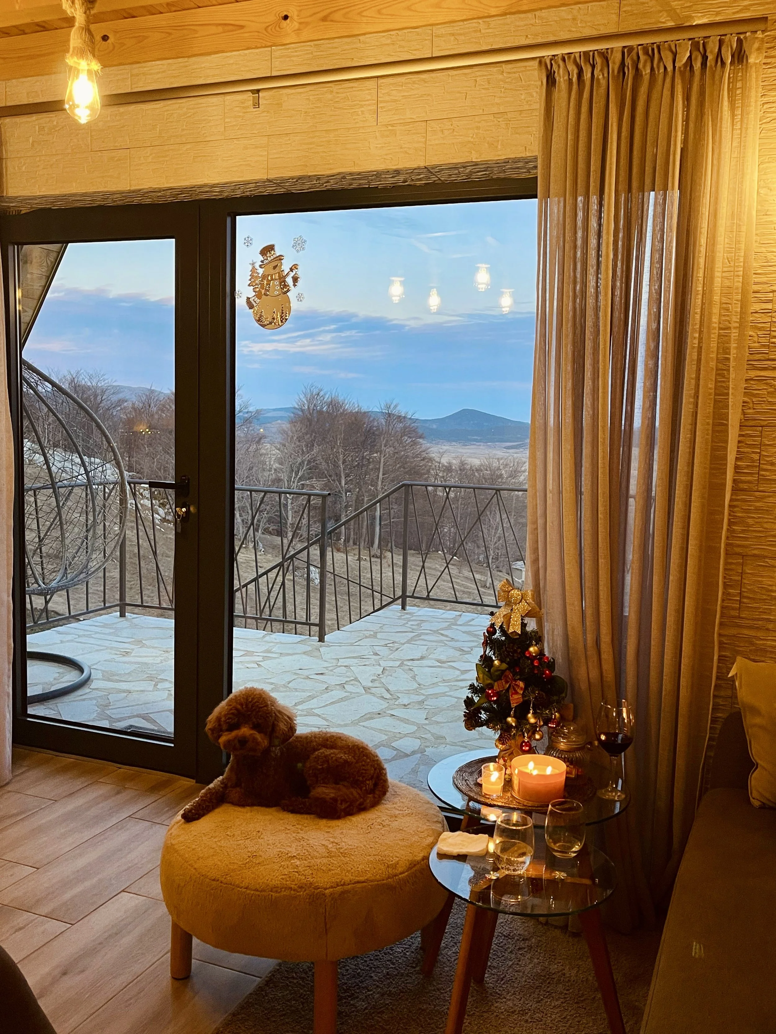 A cozy interior living room scene with a small brown dog sitting on a yellow ottoman near a glass door. The door leads to a balcony with black metal railings and a scenic outdoor view of trees, mountains, and a cloudy sky. The room is decorated with 