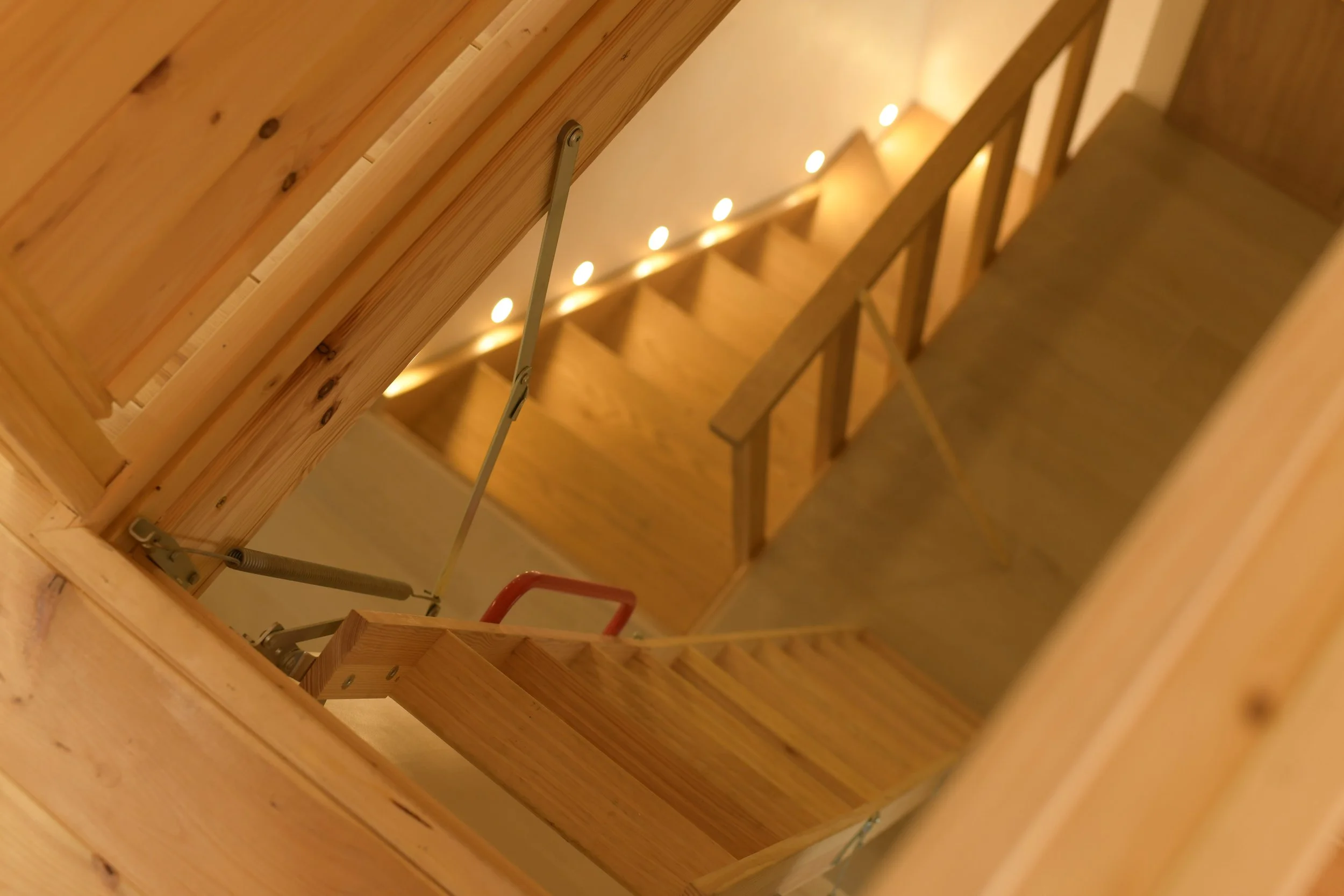 View of a wooden staircase from above, showing handrails, steps, and lighting along the wall.