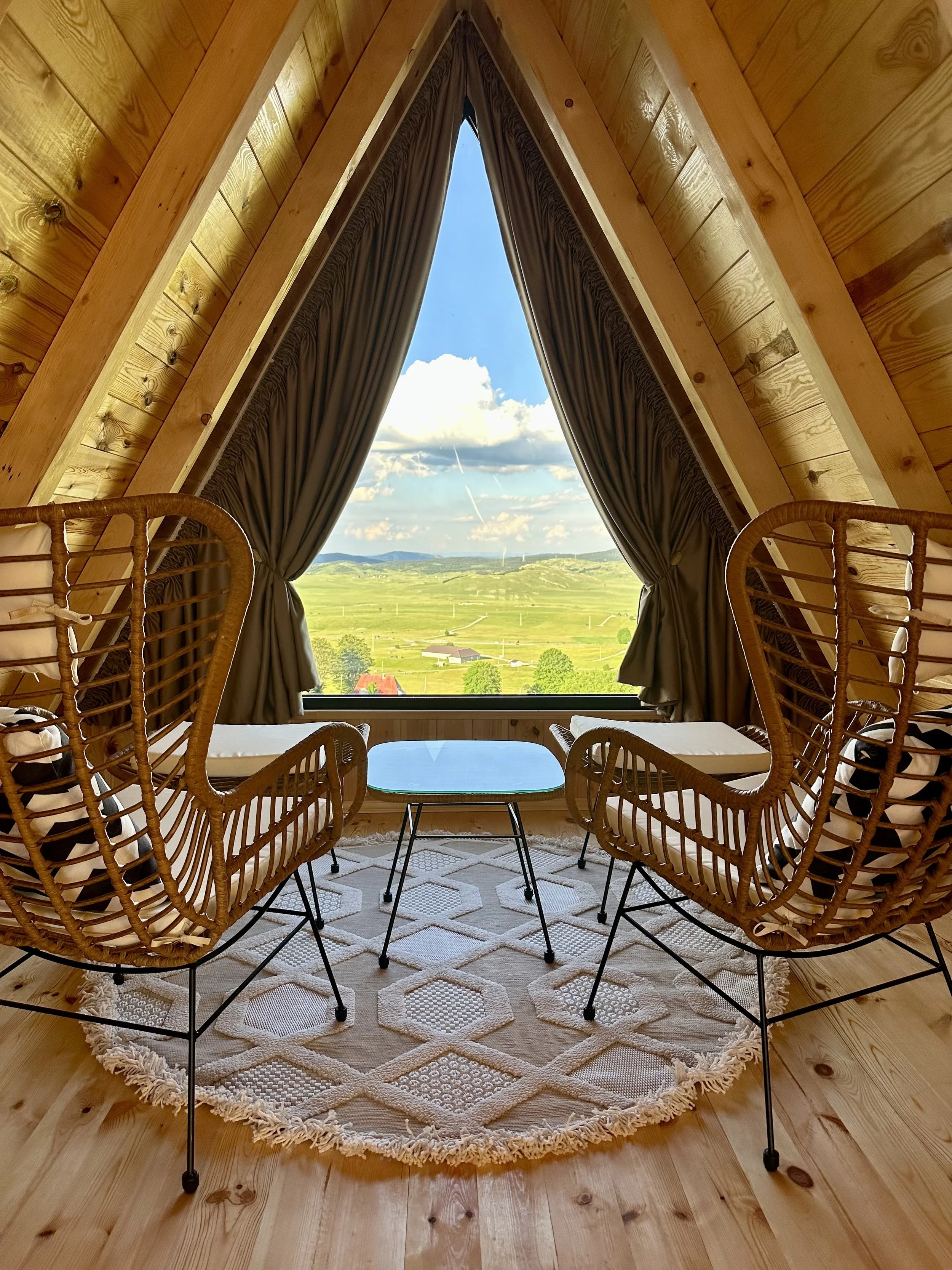Interior of a cozy room with wooden walls and ceiling, featuring a large triangular window with dark curtains, offering a scenic view of green fields, trees, and a blue sky with clouds. Two wicker chairs with cushions are positioned around a small gl