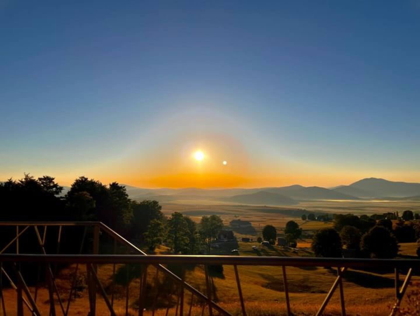 Sunset over a rural landscape with rolling hills, trees, and a wooden railing in the foreground.