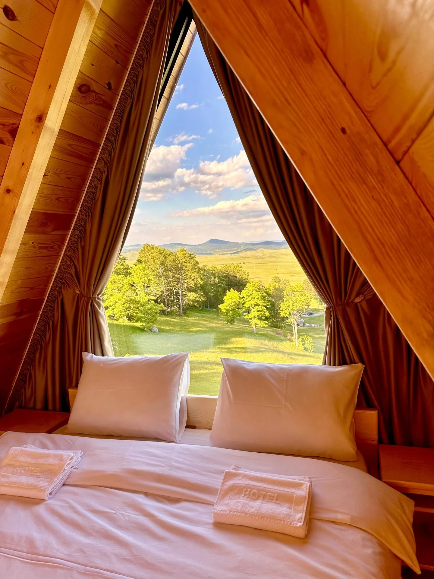 View from inside a wooden cabin with a bed, showing a large window with brown curtains, overlooking a green landscape with trees, grassy fields, distant hills, and a partly cloudy sky.