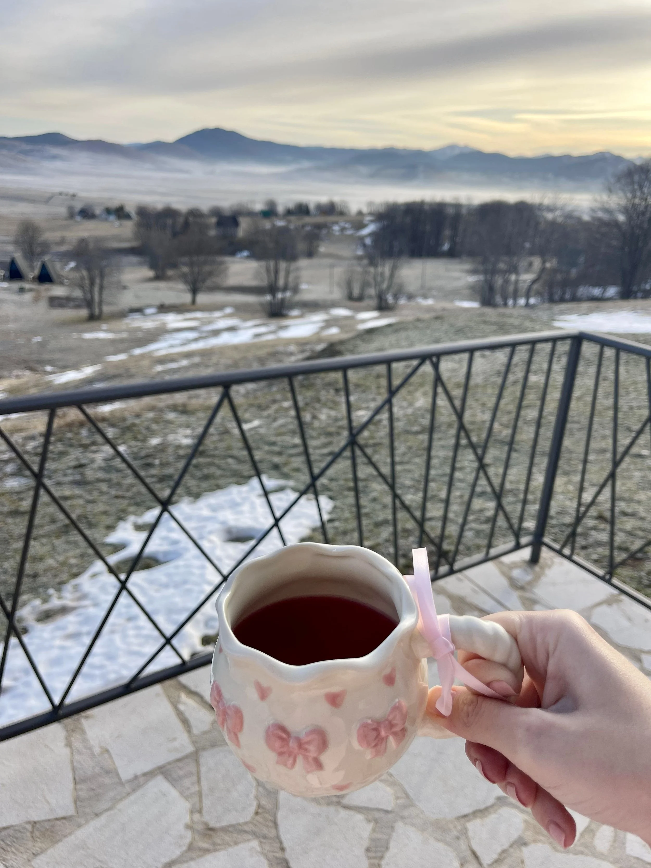 A hand holding a ceramic mug with pink bows, filled with a dark beverage, on a balcony overlooking a snowy, mountainous landscape at sunrise or sunset.