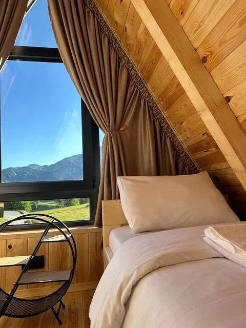 A cozy bedroom with wooden-paneled walls and ceiling, a bed with white bedding, a pillow, and a folded towel. There's a large window with beige curtains, showing a mountain view outside.