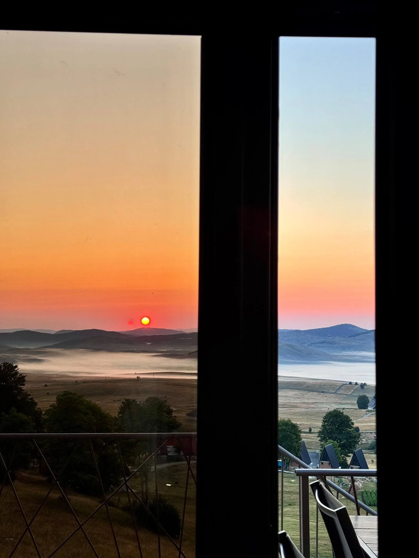 View of sunrise over mountains seen through a window with a sliding glass door, with a balcony and trees in the foreground.