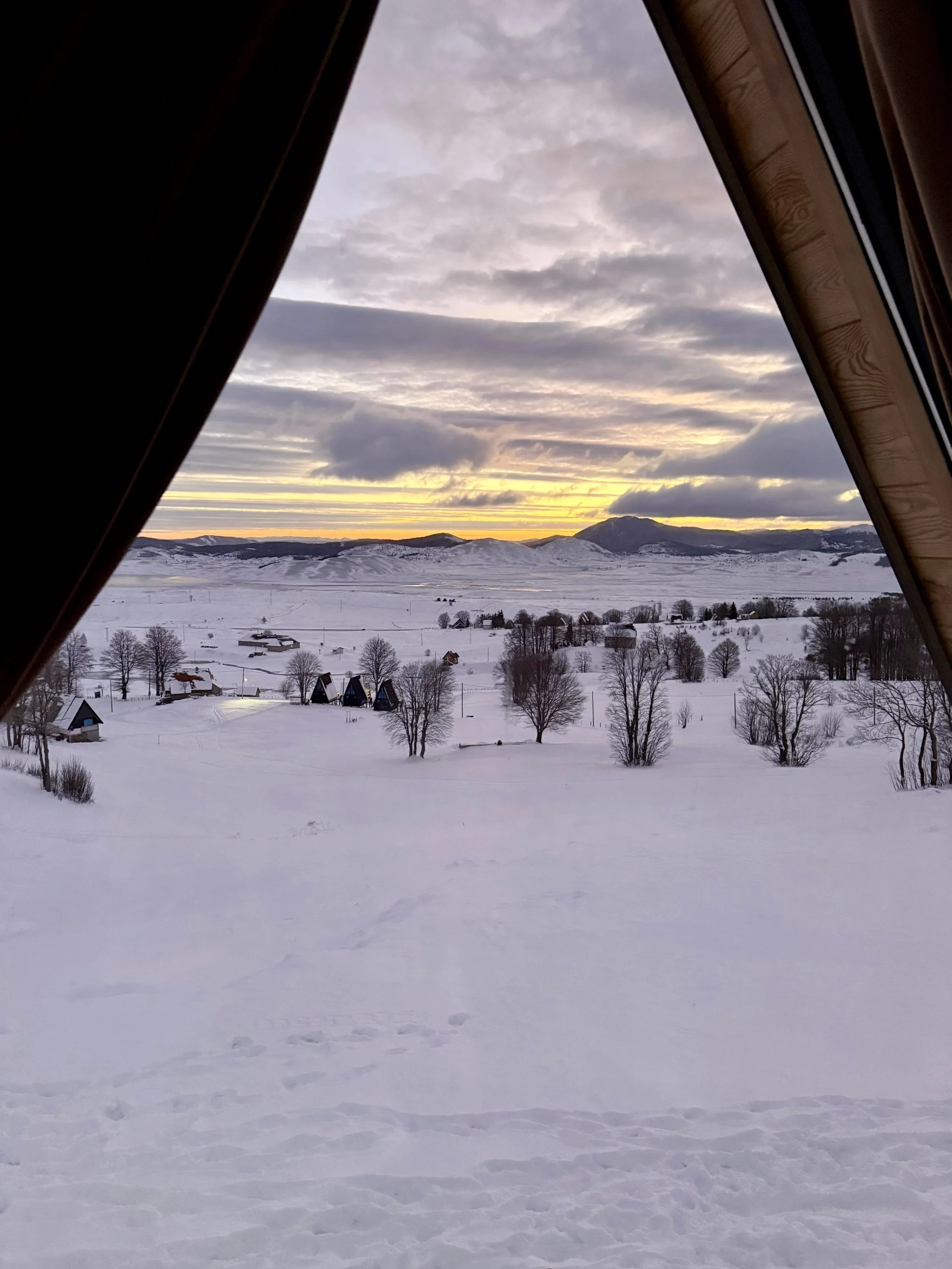 Snow-covered landscape viewed from a window at sunset, with small houses, trees, and distant mountains under a cloudy sky.