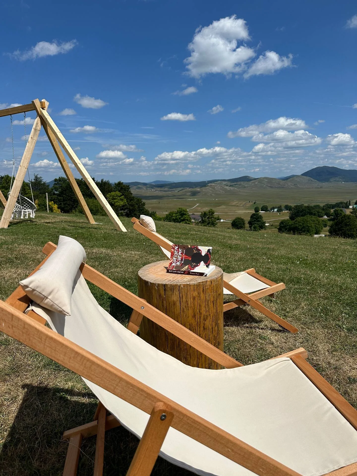 Two white deck chairs with wooden frames on a grassy hill, facing a scenic landscape of rolling hills and mountains under a blue sky with white clouds. A small round wooden table with a book on top is between the chairs, and a wooden swing set is in 