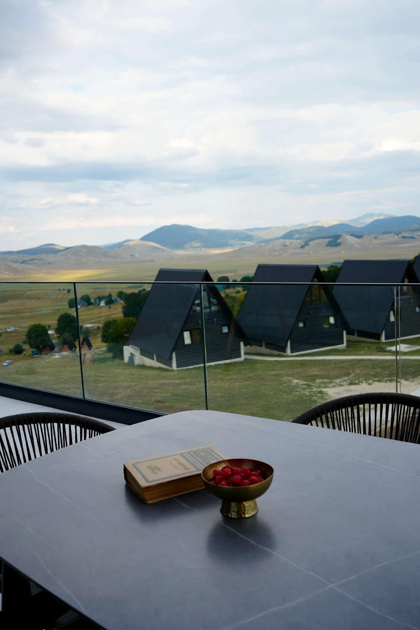 A view from a balcony with black A-frame houses in a rural landscape, mountains in the background, and a table with a bowl of raspberries and a menu or booklet.