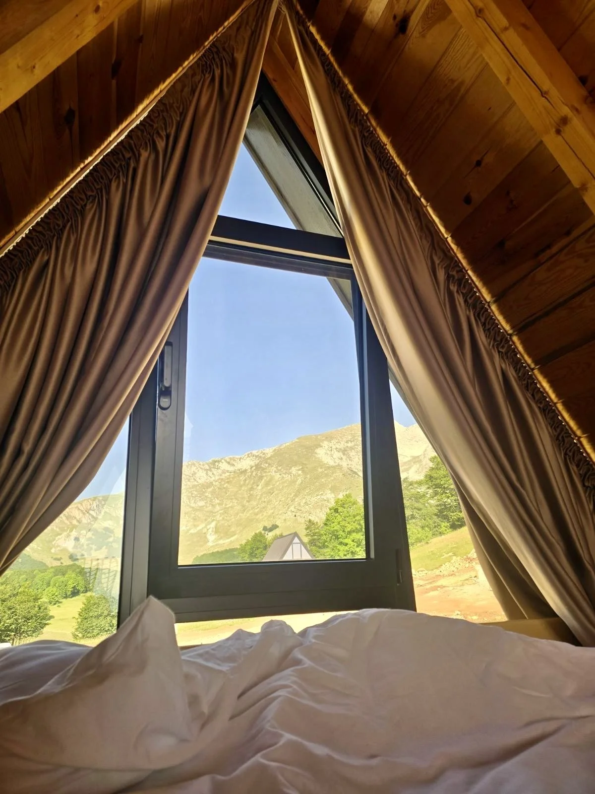 View of a mountain landscape outside a window with beige curtains inside a wooden cabin, including part of a bed with white sheets in the foreground.