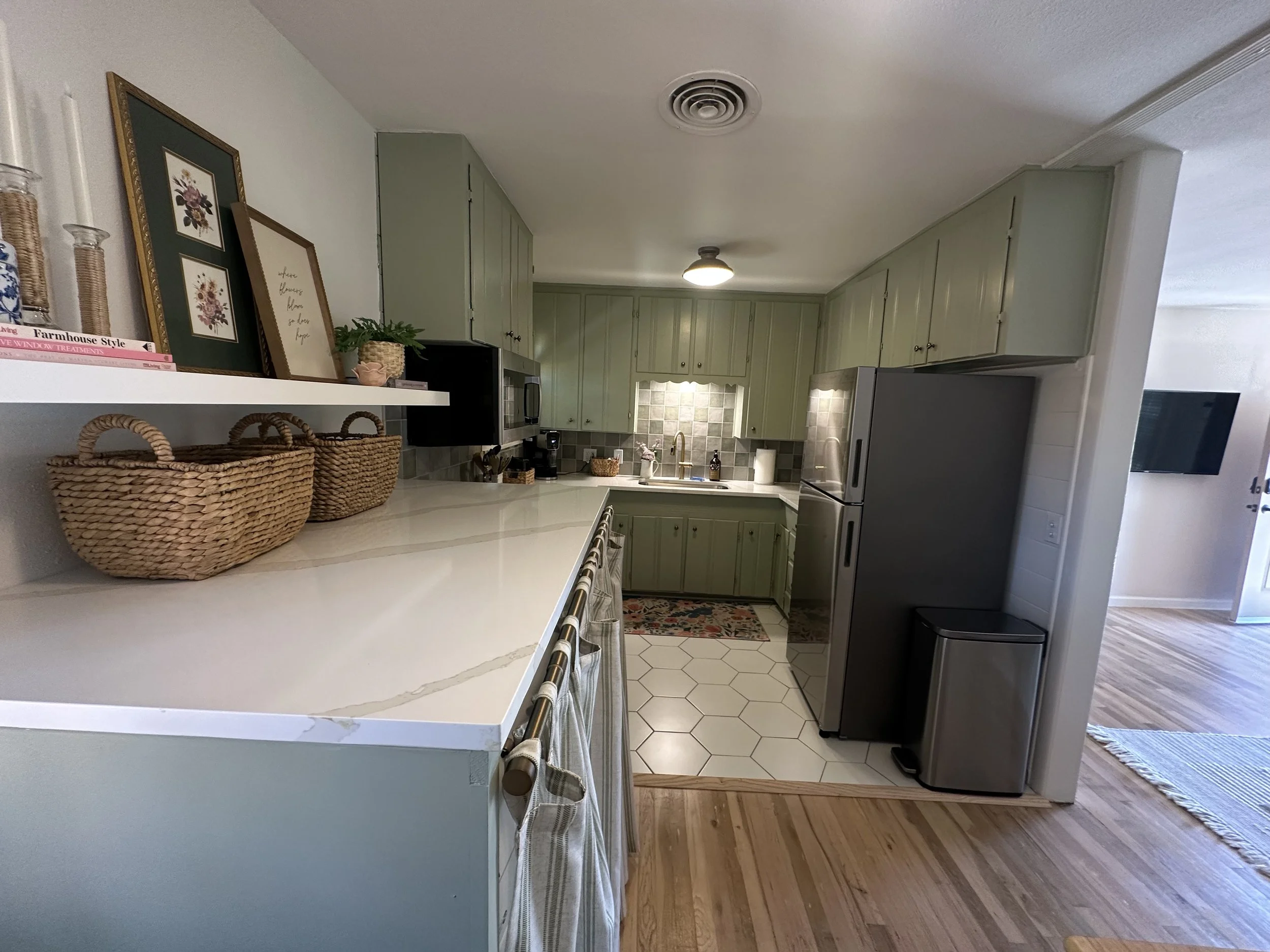 A kitchen with light green cabinets, a gray refrigerator, a white countertop, and a small tiled floor area. Decor includes framed artwork, woven baskets, and a small potted plant.