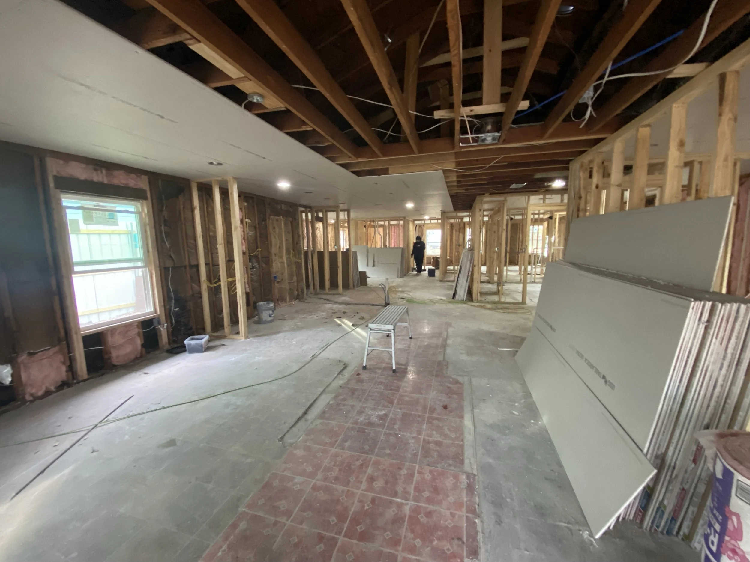 Interior view of a house under renovation with exposed wooden framing, construction materials, and debris on the unfinished floor. There is a window on the left wall and a person working in the background.