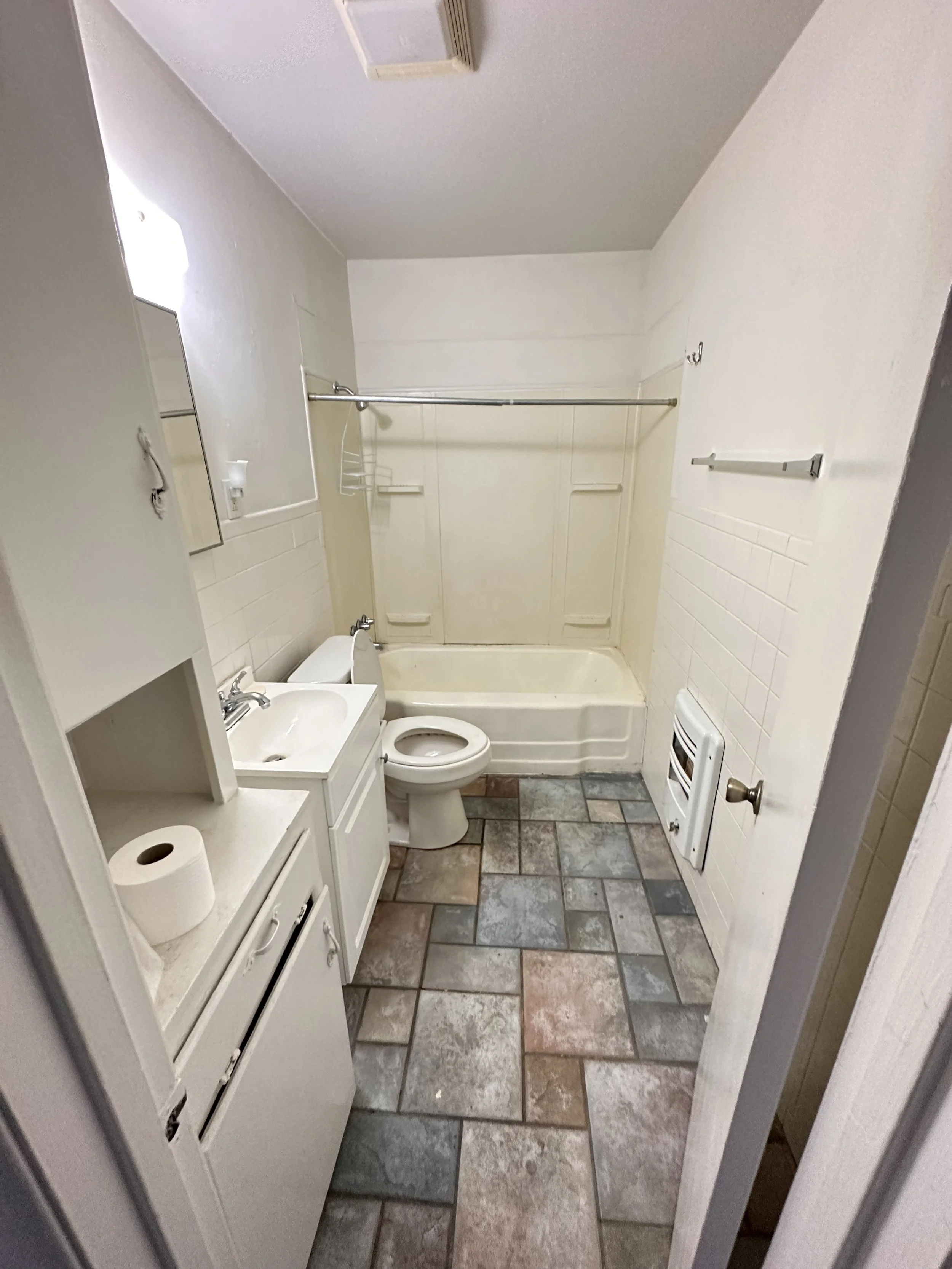 Small bathroom with tiled floor, white sink with cabinet, toilet, and a bathtub with a shower curtain rod. There is a wall heater or vent near the floor and a mirror above the sink.