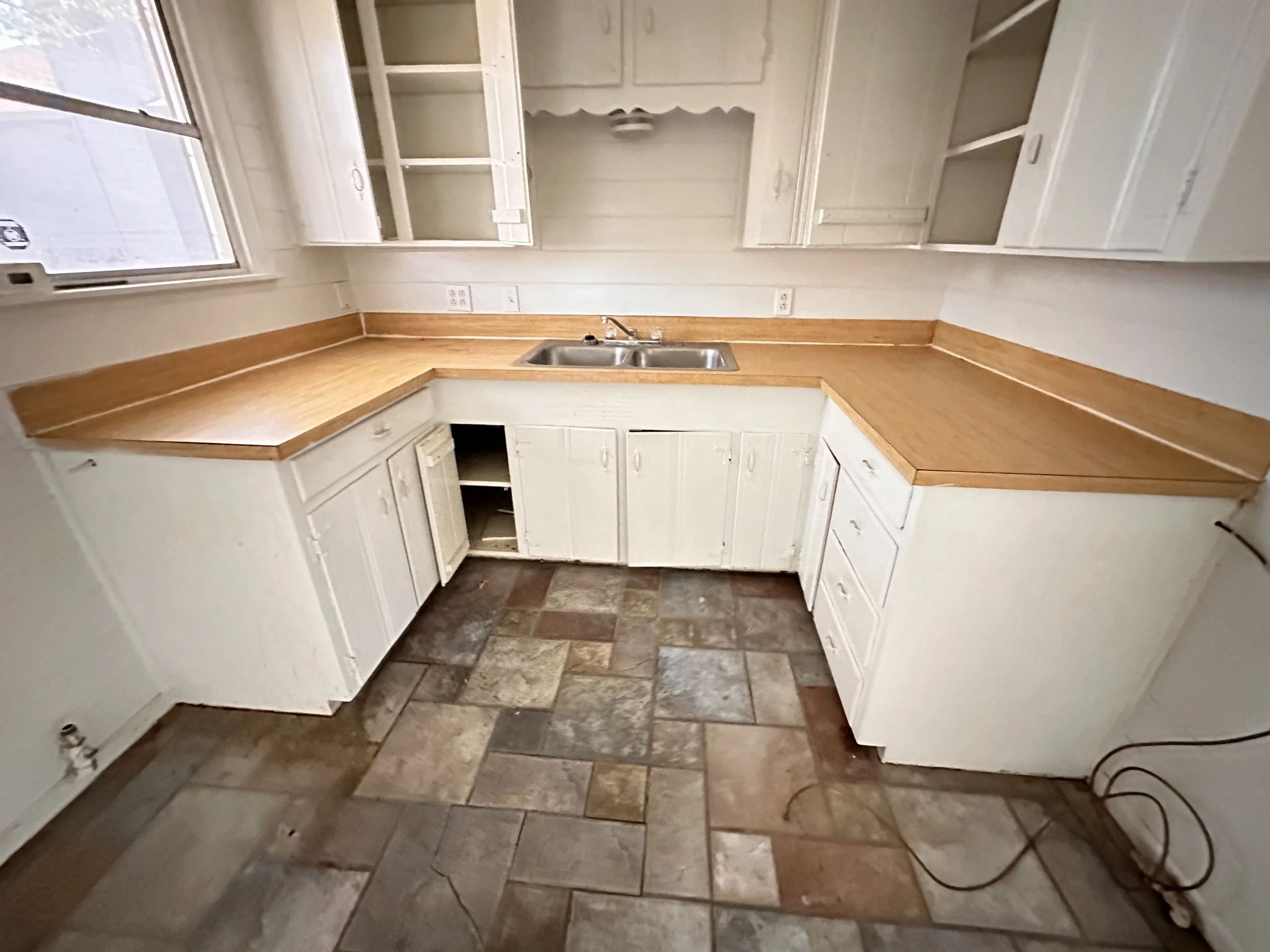 Empty kitchen with white cabinets, a wooden countertop, a double sink, and a window, with tiled flooring.