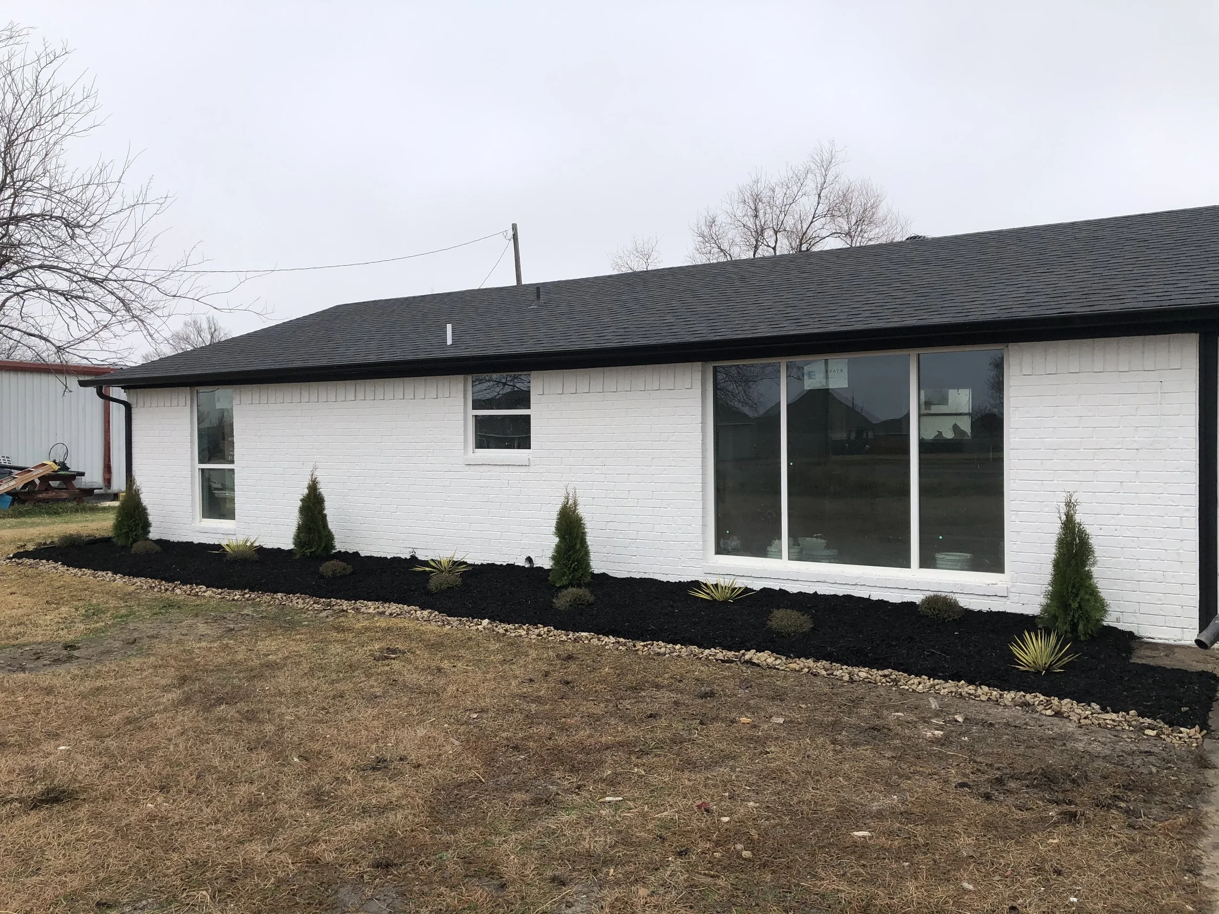 A white brick house with large windows and a black roof, surrounded by a freshly landscaped garden bed with small bushes and mulch.