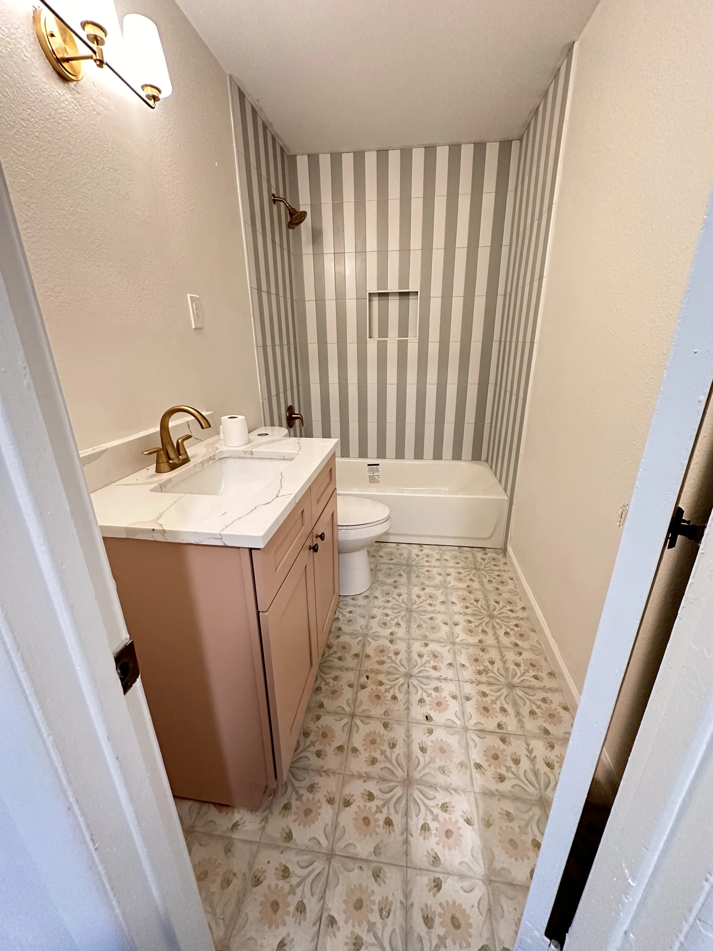 A small bathroom with a pink vanity with a marble countertop, a brass faucet, and a toilet next to it. The shower area has striped beige and gray tiles with a built-in shelf, a brass showerhead, and a white bathtub. The floor has floral-patterned til