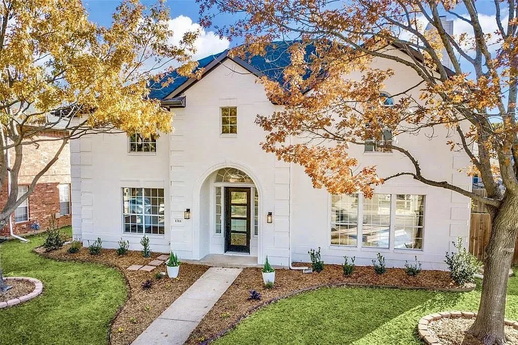 White two-story house with black door, large front windows, surrounded by green lawn, orange and yellow fall trees, and a curved concrete walkway.