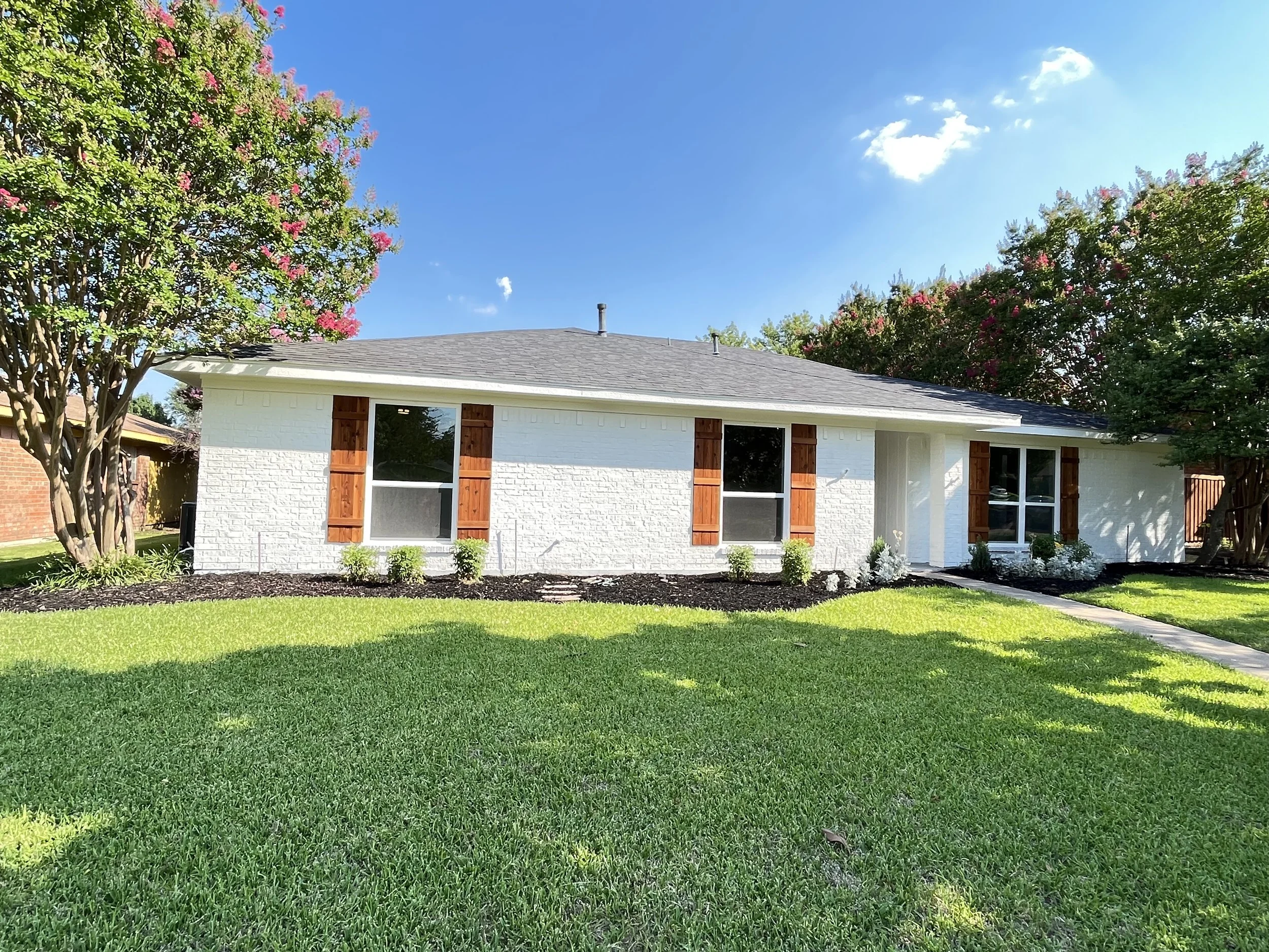 A single-story white house with brown shutters, a gray roof, and a small front porch. The house is surrounded by a green lawn with trees and flowering bushes, under a bright blue sky with a few clouds.