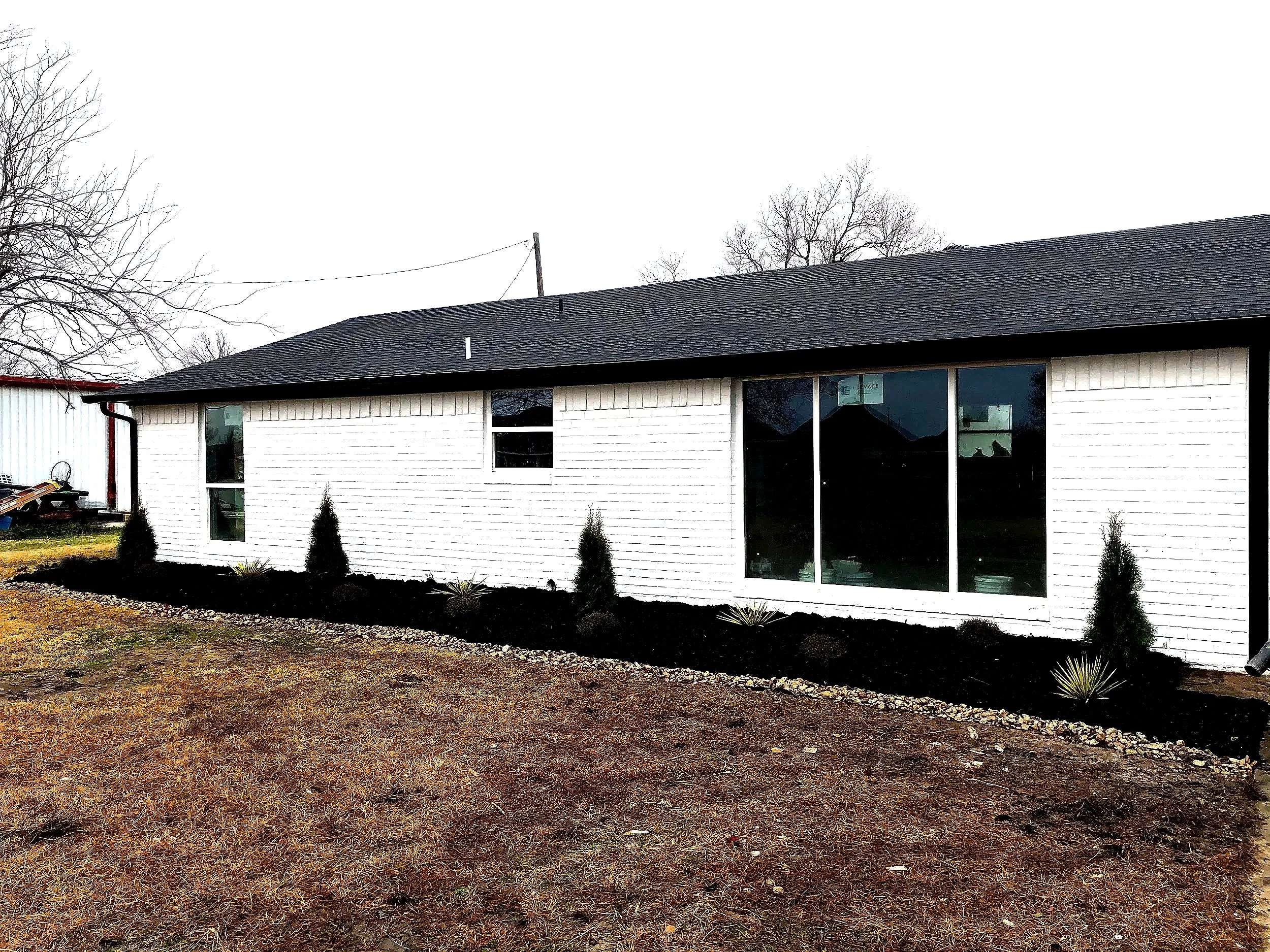 Front view of a house with white brick exterior, black roof, large sliding glass door, and small shrubbery in the garden bed.