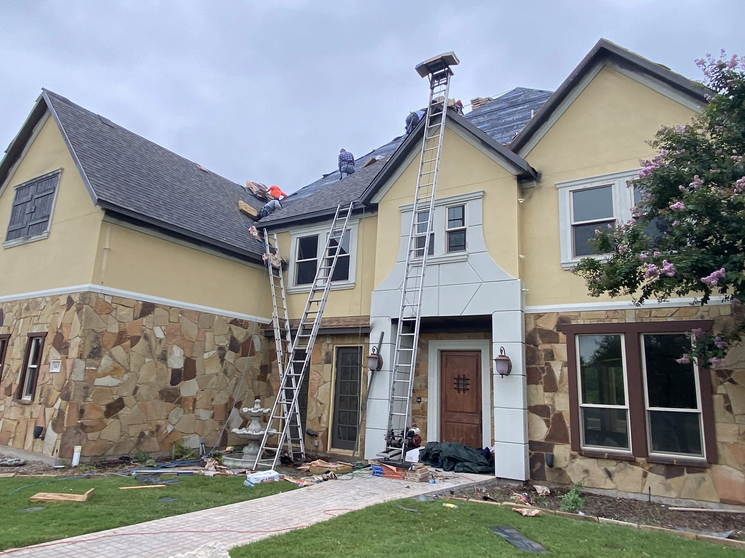 Workers repairing or replacing the roof of a two-story, yellow and stone house using ladders and safety equipment.