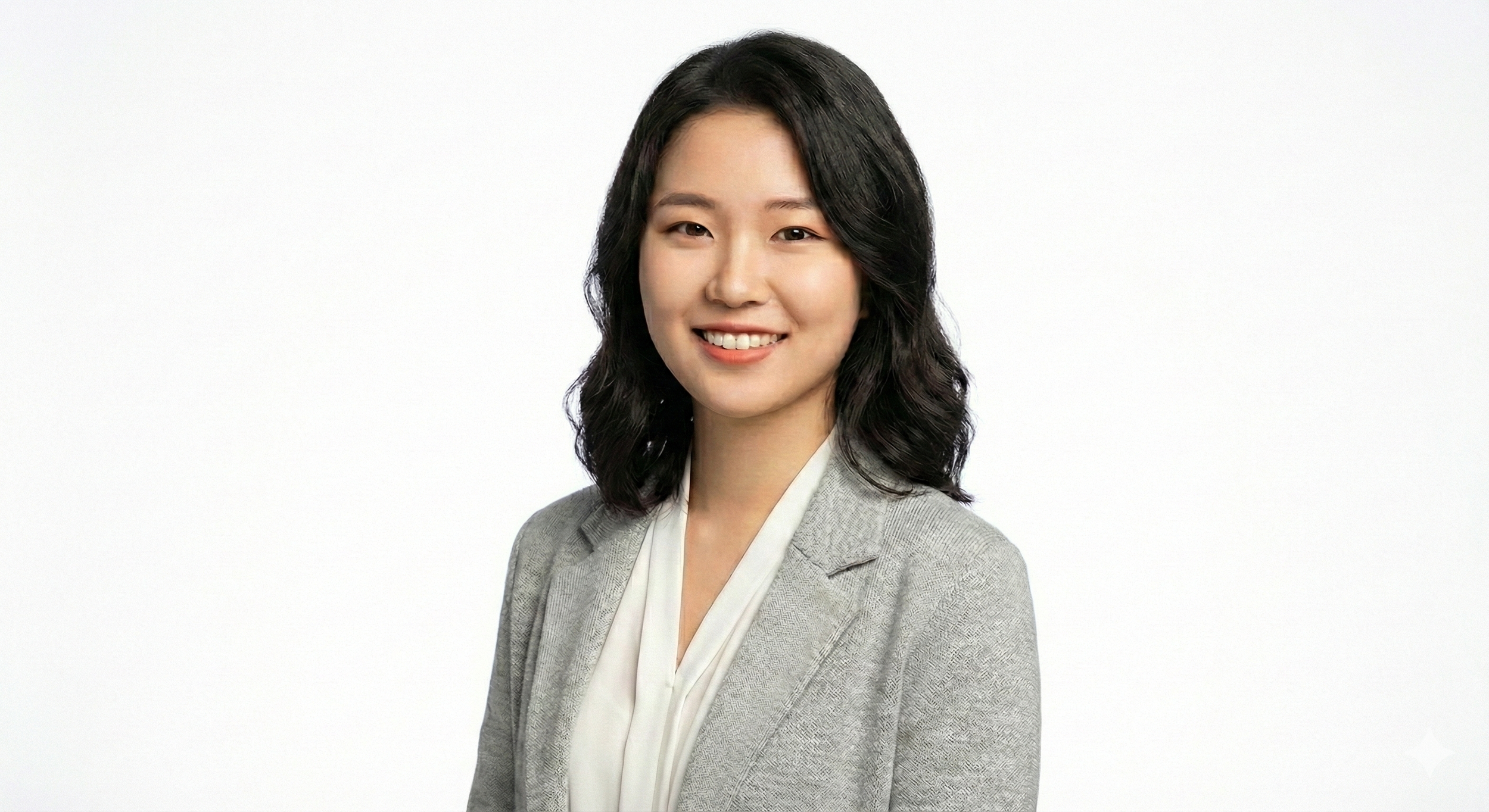 Professional woman with black wavy hair smiling, wearing a light gray blazer and white blouse, against a plain white background.