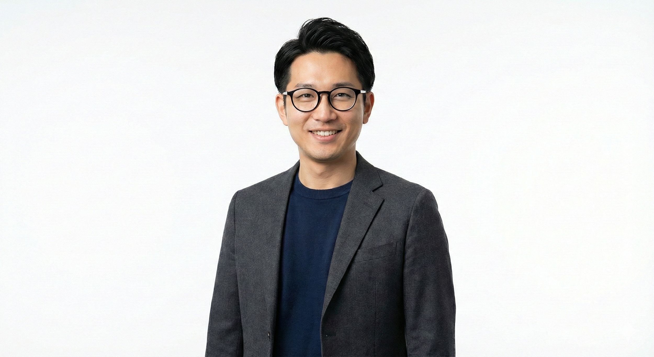 Man with black hair, glasses, wearing a dark blazer over a navy shirt, smiling, against a plain white background.