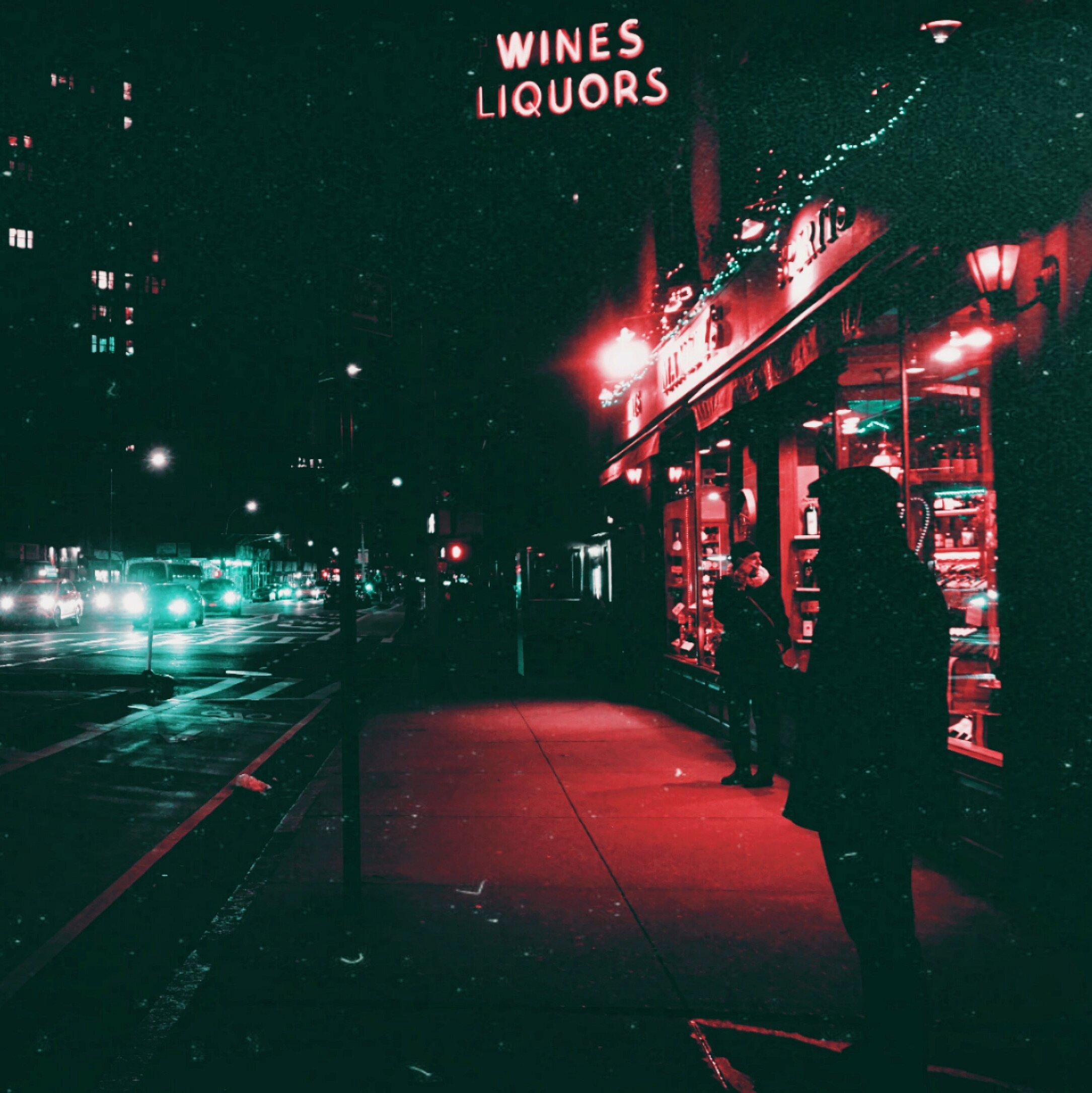 Nighttime street scene with a lit wine and liquor shop, two people standing outside, and cars with headlights on driving along the street.