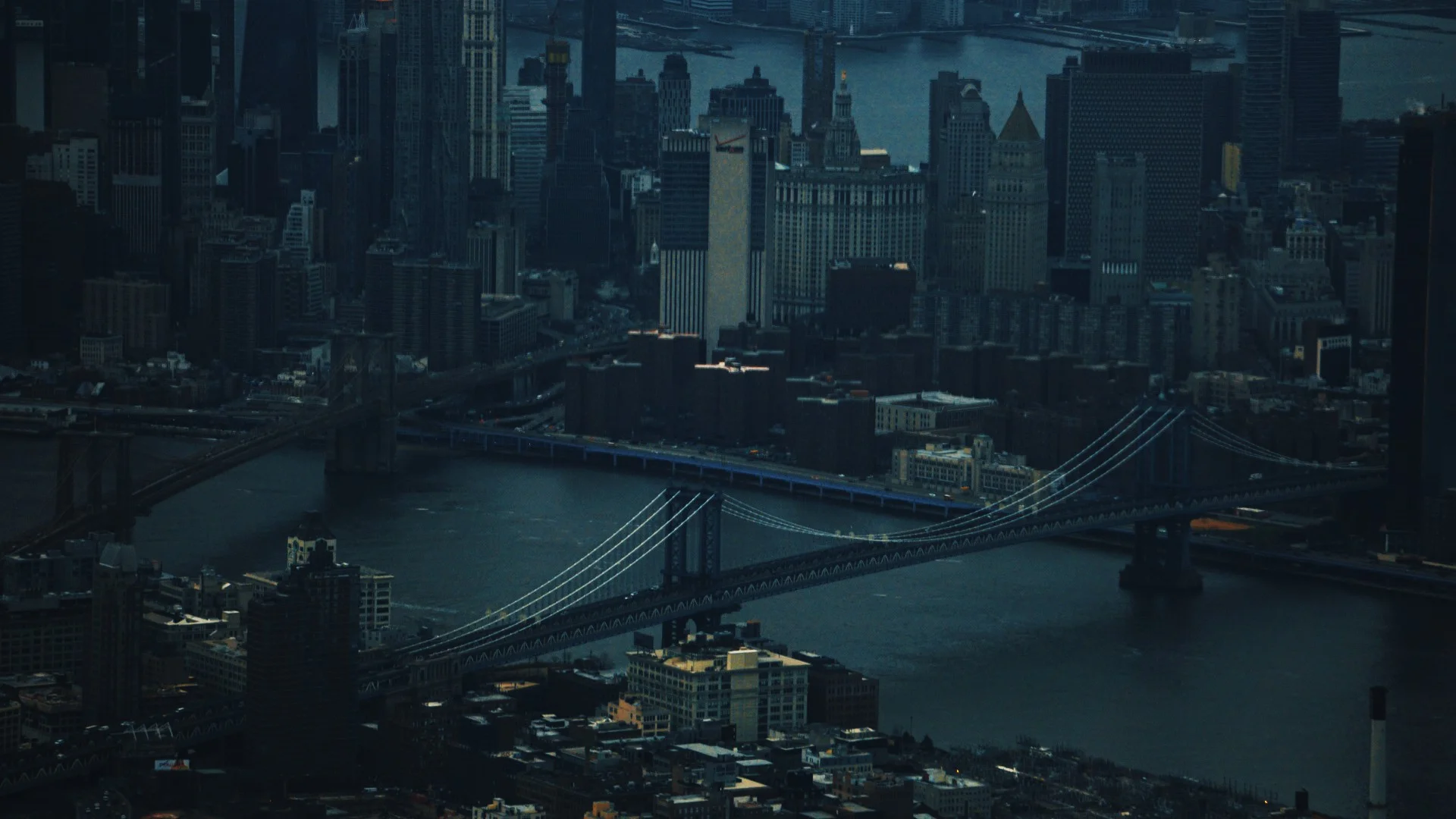 An aerial view of a large suspension bridge spanning a river connected to a dense skyline of skyscrapers in low light.