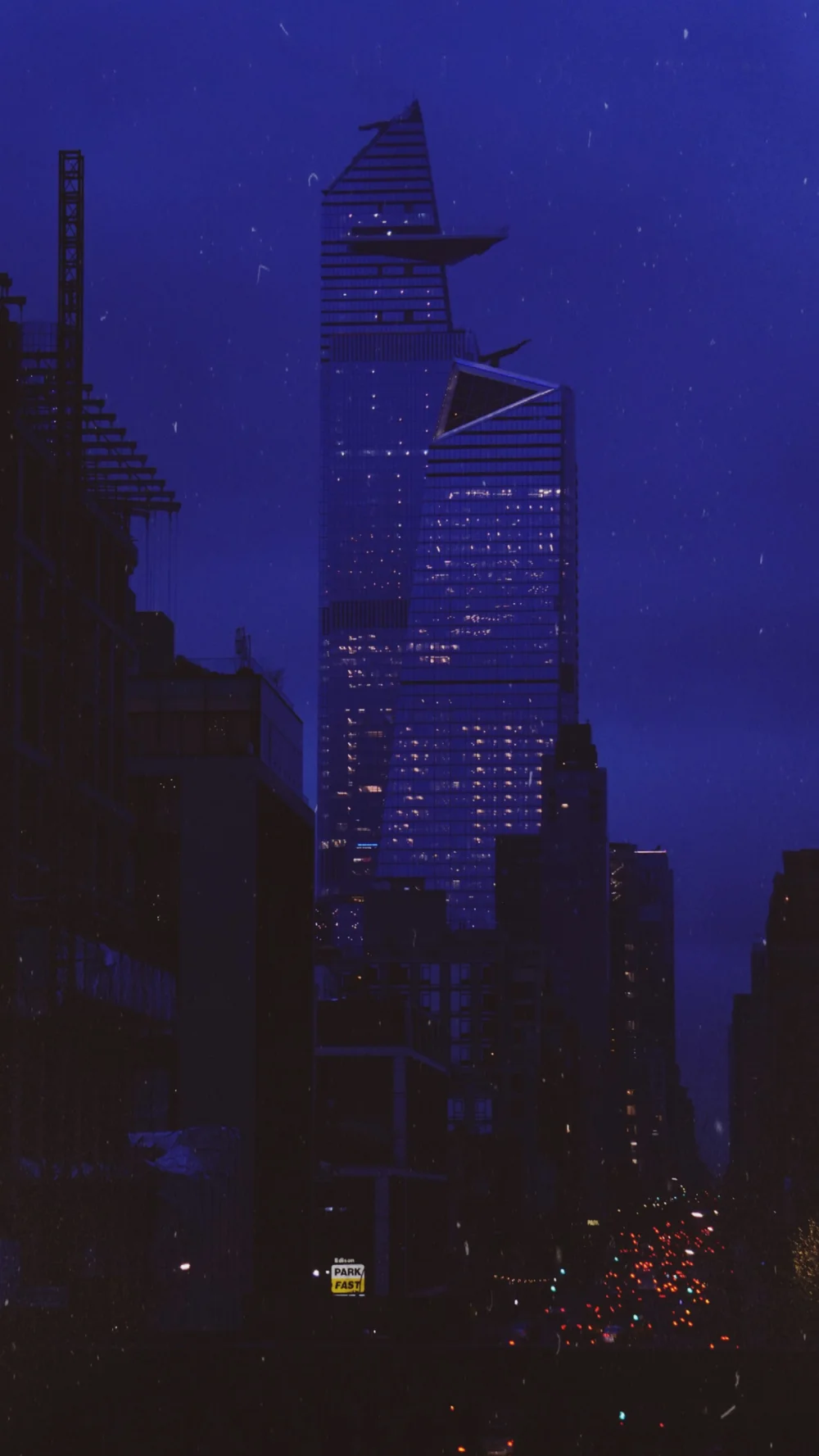 A skyscraper at night between darker buildings, under a deep blue sky.