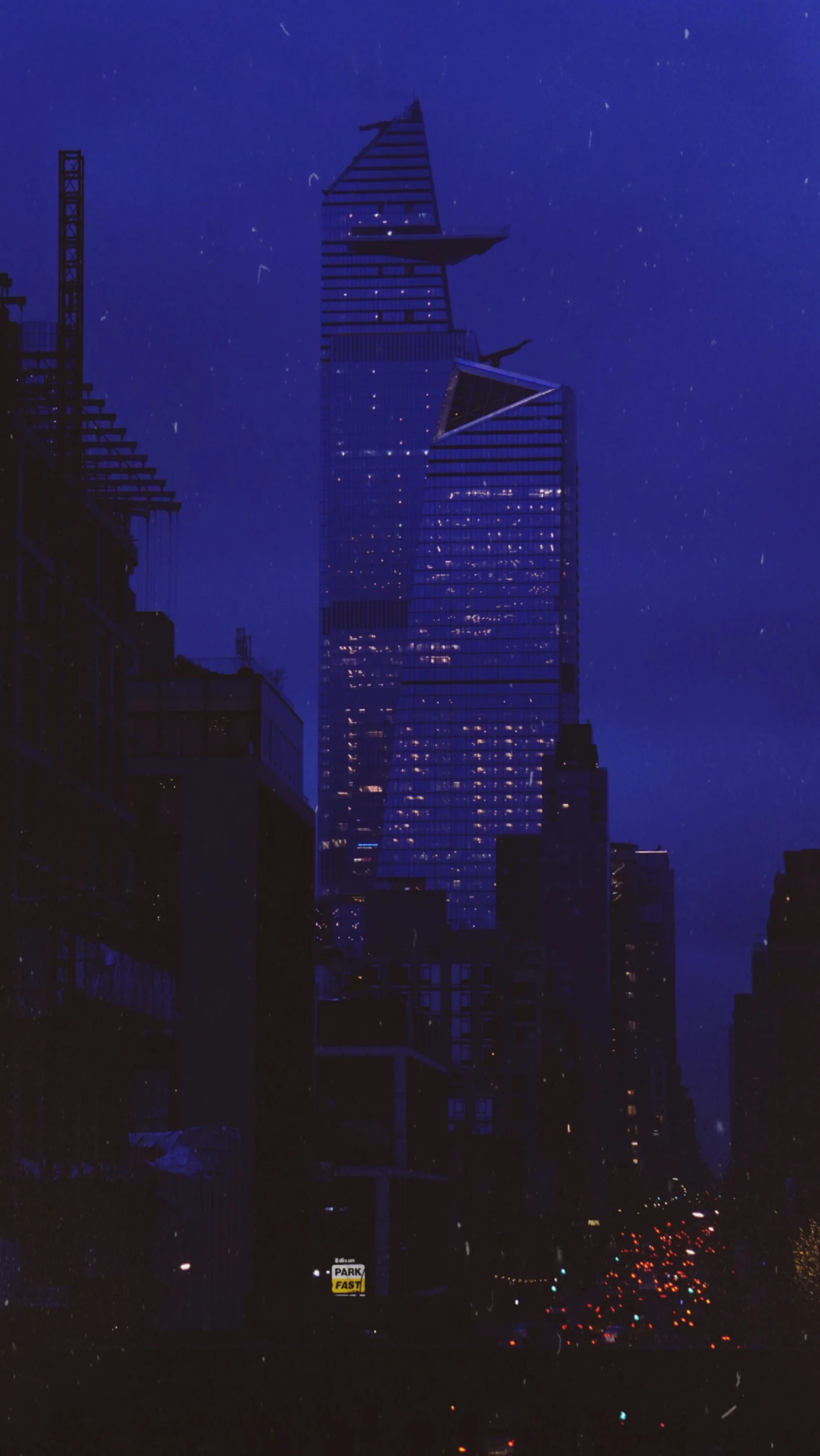 A skyscraper at night between darker buildings, under a deep blue sky.