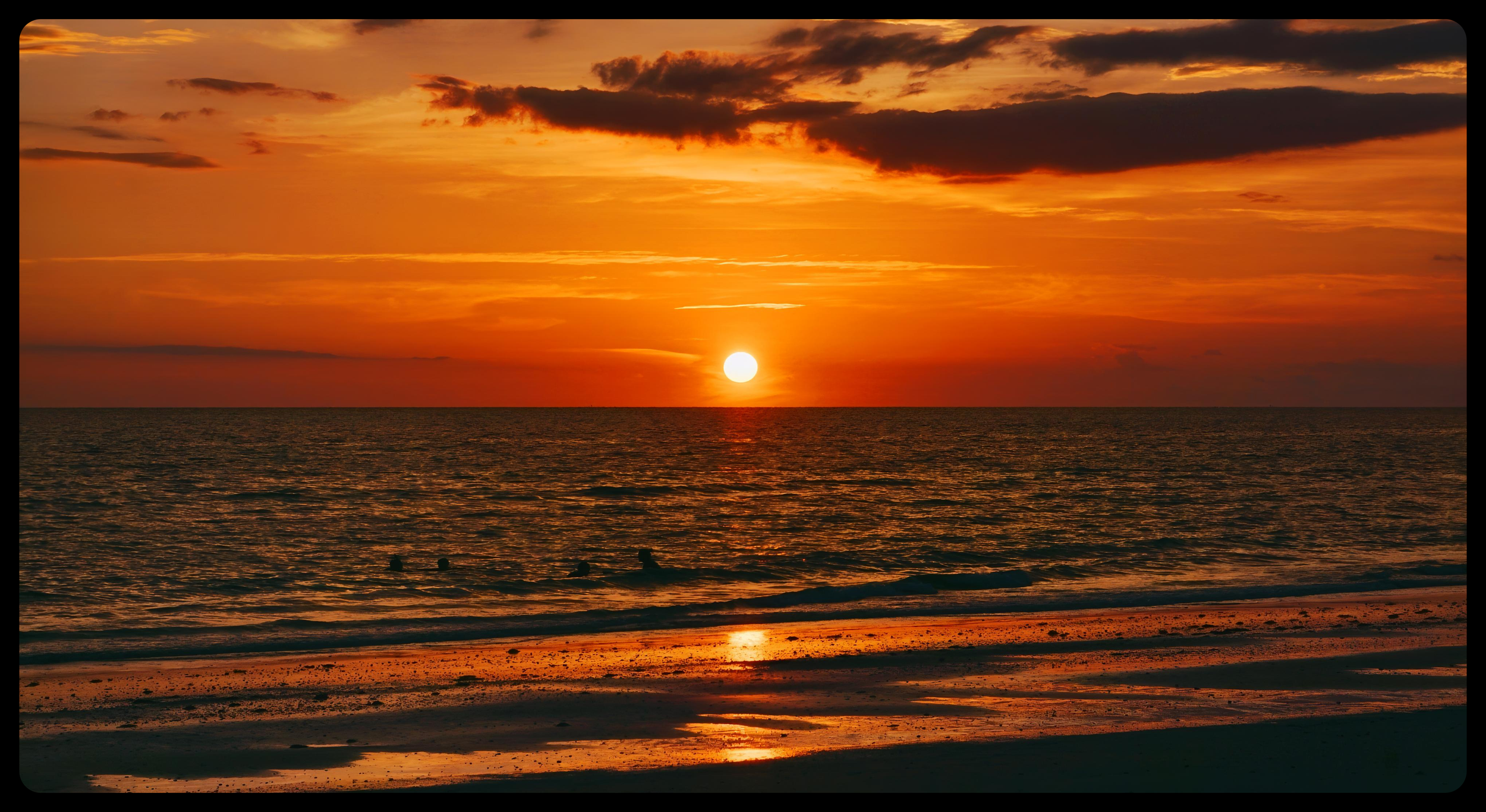 A vibrant sunset over the ocean with the sun low on the horizon. The sky features orange, yellow, and dark clouds. The water reflects the colors of the sunset, with gentle waves and some silhouetted figures swimming in the distance.