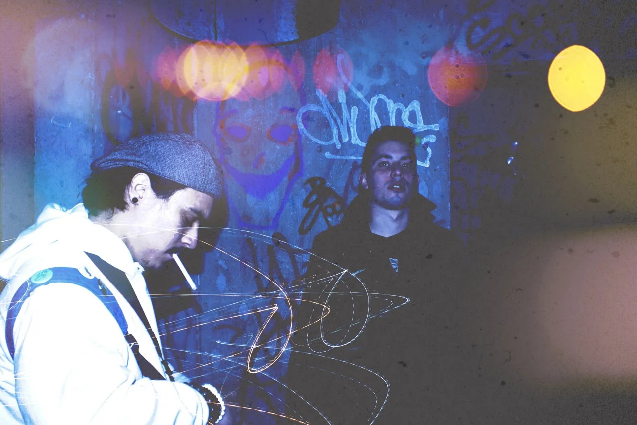 Two young men standing against a graffiti-covered wall at night. One is wearing a gray beanie and white jacket, looking down with a cigarette in mouth, while the other is facing the camera with a dark jacket, illuminated by colorful lights.