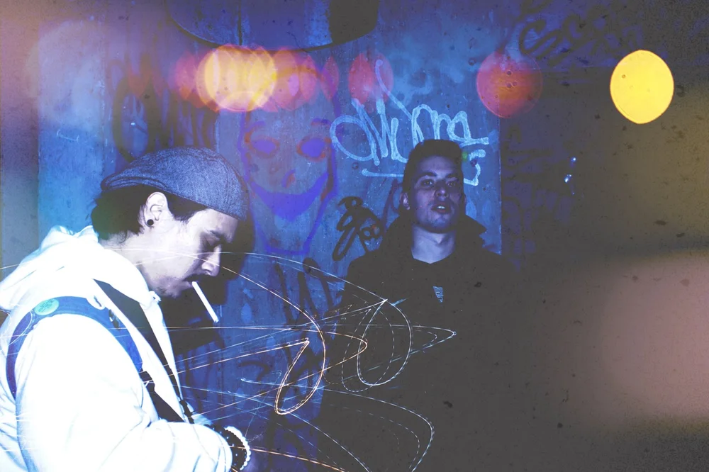 Two young men by a graffiti wall at night, one smoking, with light trails.