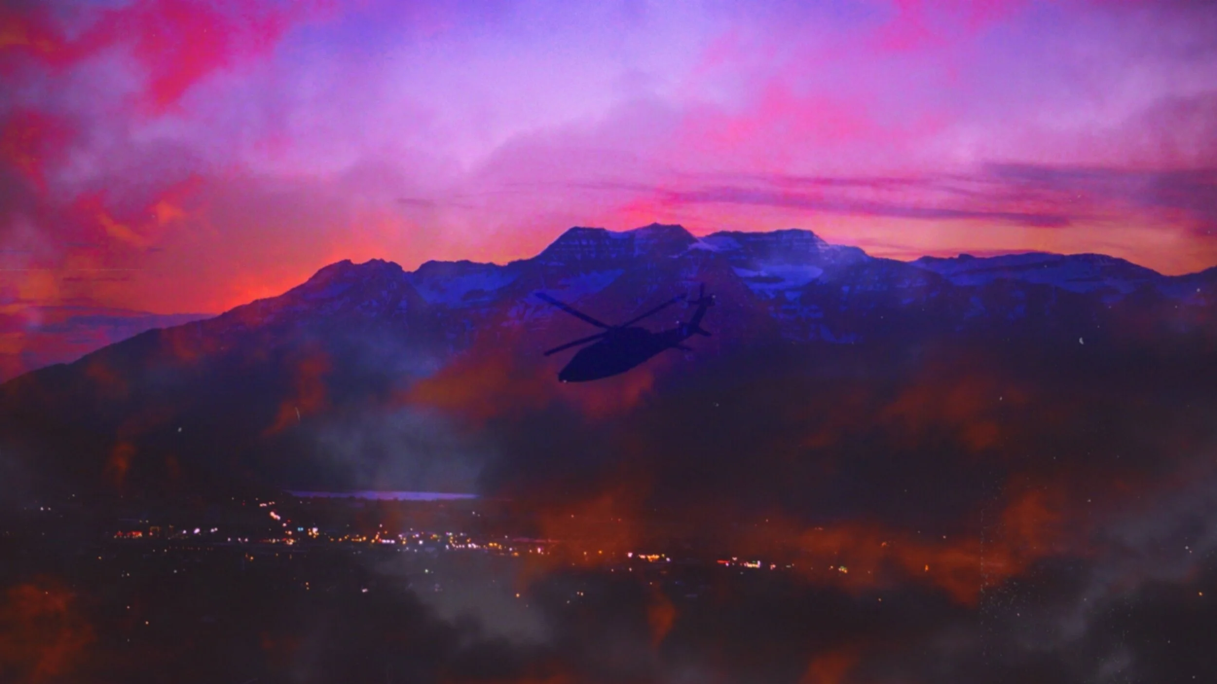 A helicopter flying near a mountain range during a colorful sunset with pink, purple, and orange hues, and smoke or fog near the ground.