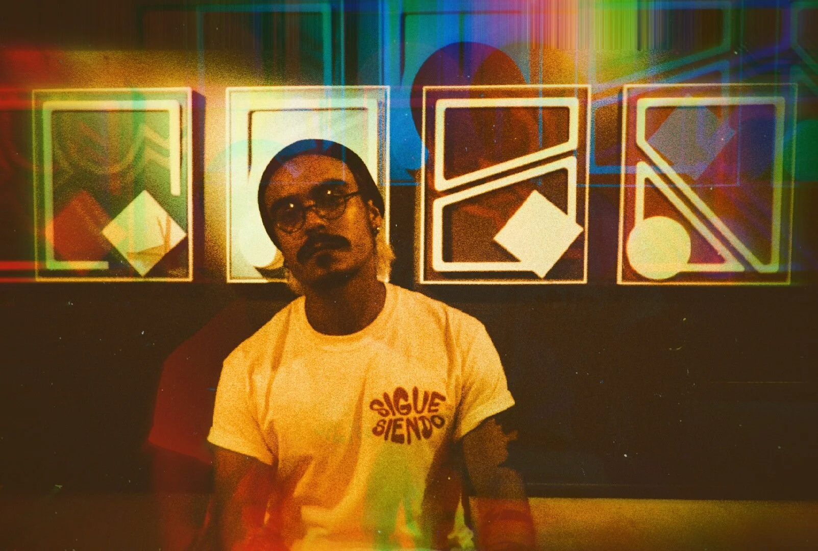 A man in glasses and a printed T-shirt seated in front of neon geometric artwork.