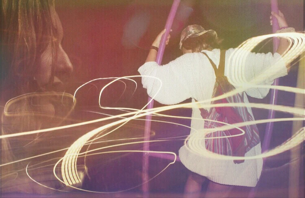 A person holding a pole or stick, moving it in an environment with light trails, and a faded image of a woman's face in the background.