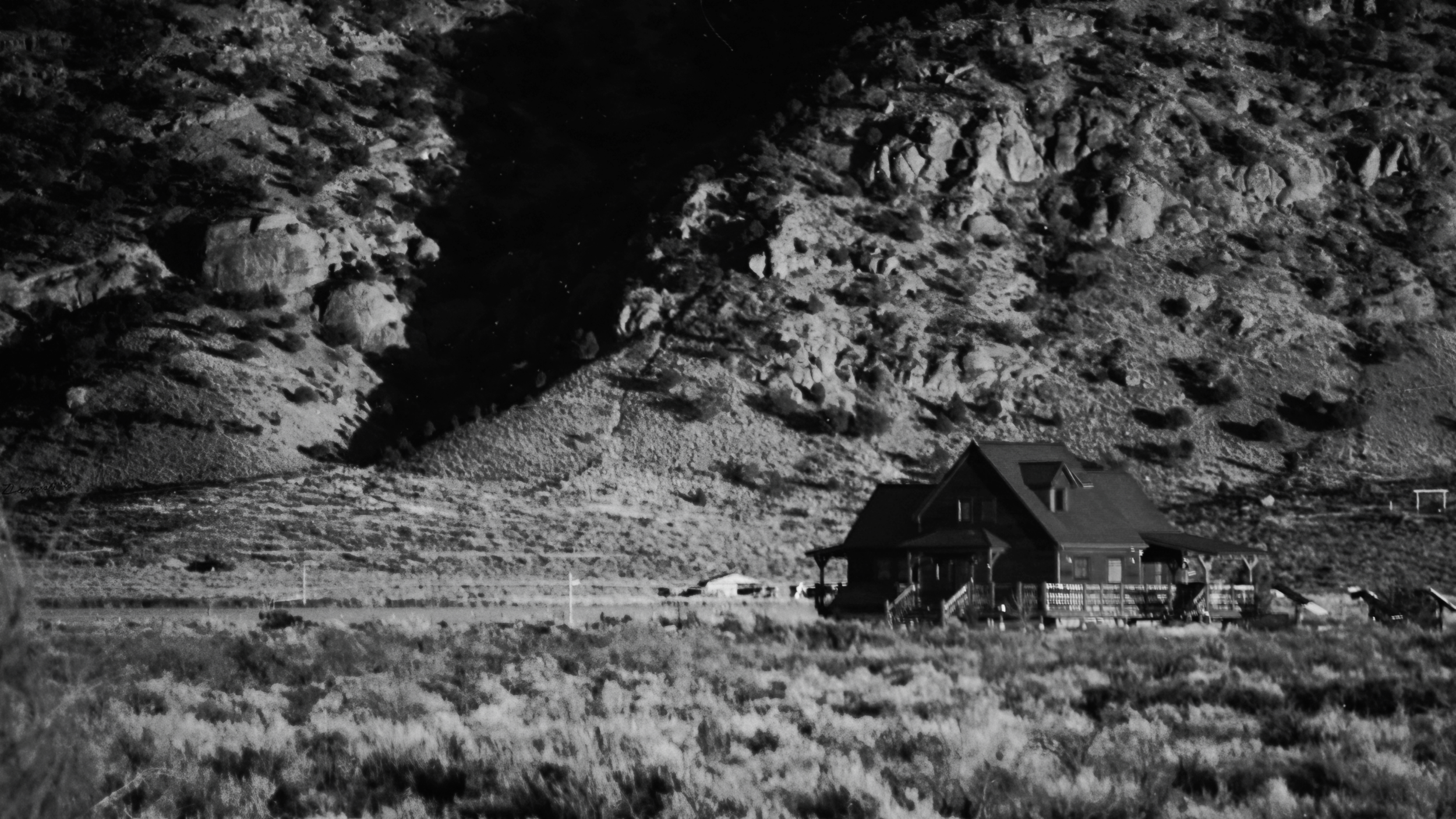 A solitary wooden cabin situated in a field of brush at the base of a steep, rocky hillside, depicted in grayscale.