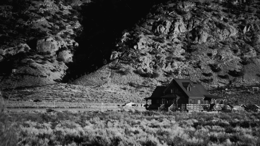 Black and white photo of a small cabin at the base of a rocky hillside.