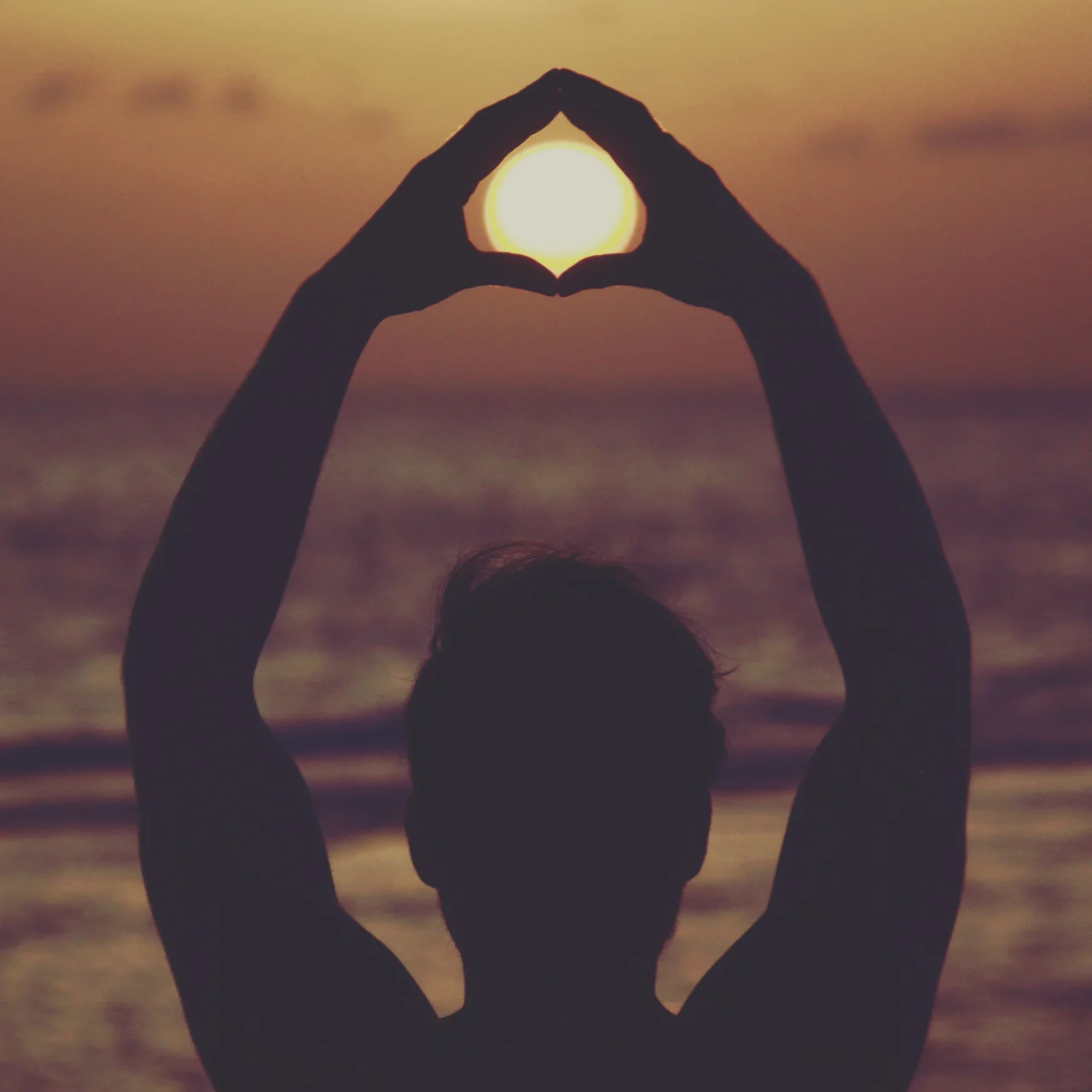 A silhouette of a person at the beach making a heart shape with their hands around the setting sun during sunset.