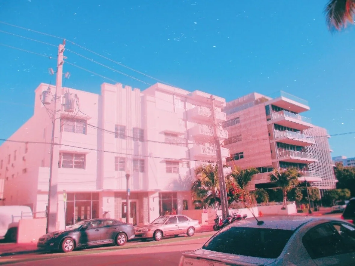 Pink-tinted building on a city street with parked cars, palm trees, and a utility pole against a clear blue sky.