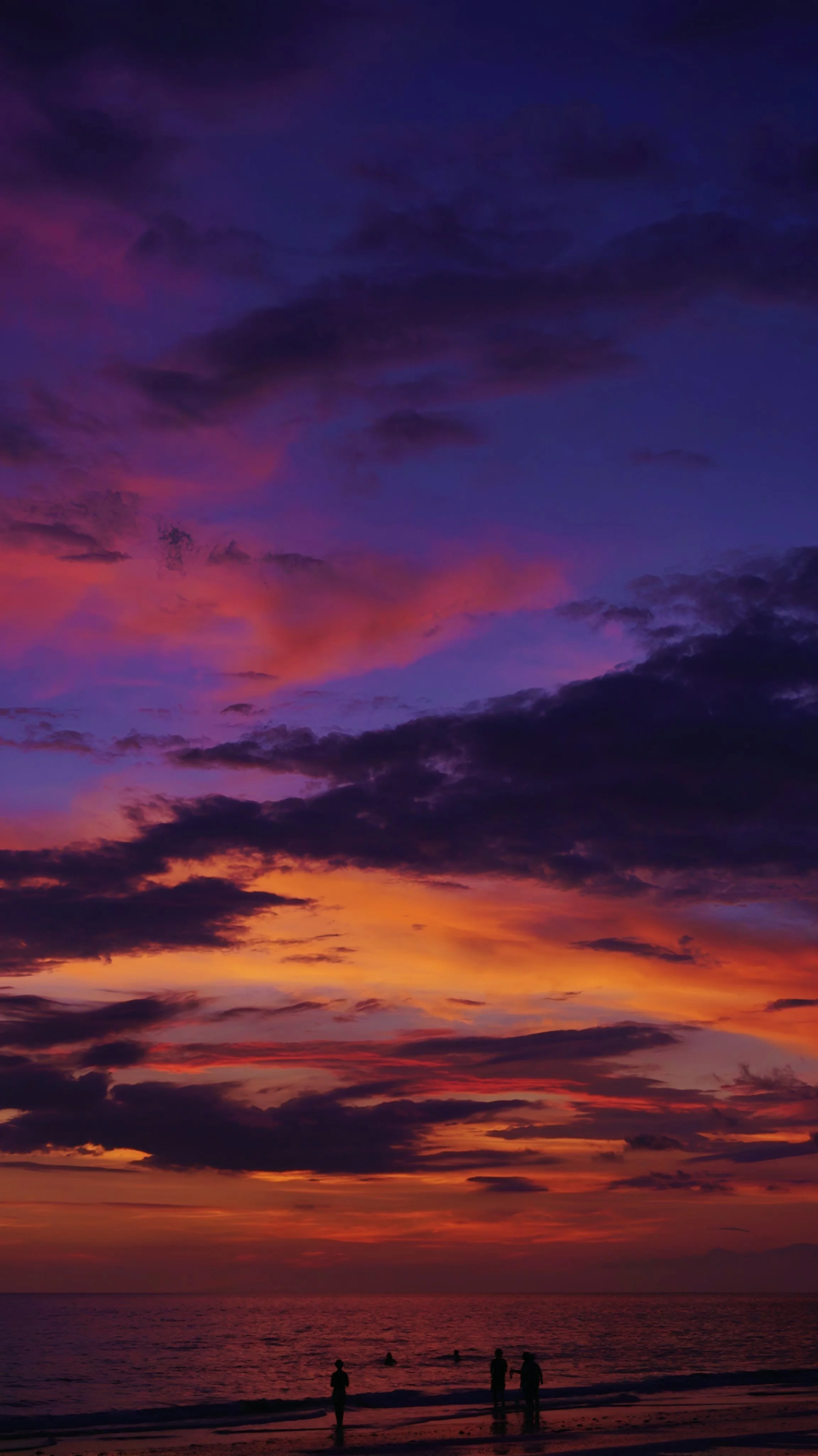 People watching the colorful sunset on the beach with purple, pink, orange, and yellow clouds.
