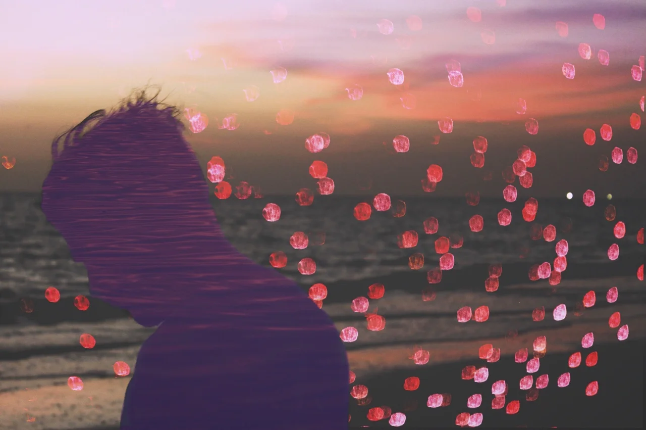 Silhouette of a person with windswept hair facing a sunset on the beach with pink and orange sky, and an overlay of pink bokeh lights.