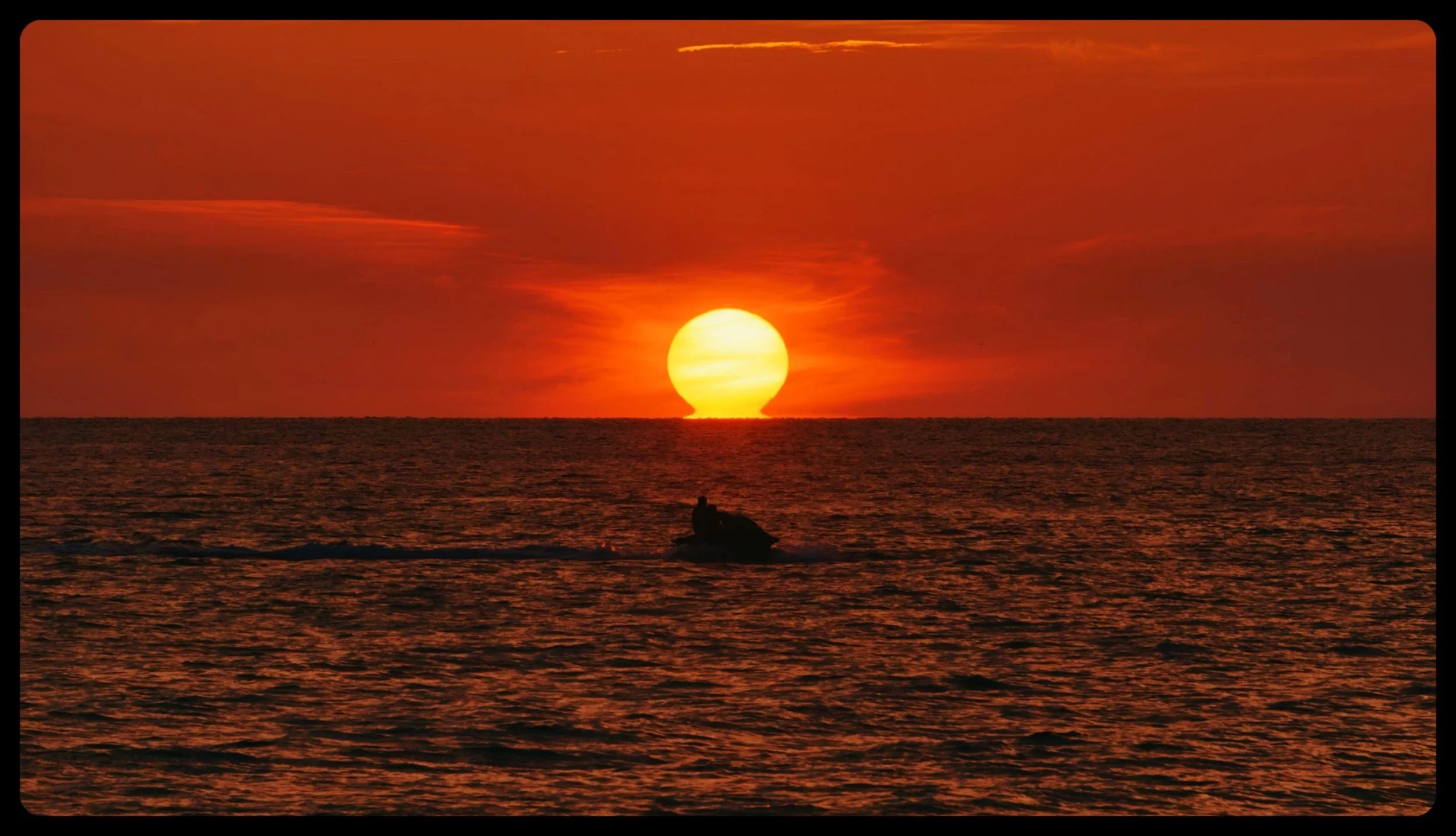 A sunset over the ocean with an orange sky, a large sun on the horizon, and a jet ski with a person sailing across the water.