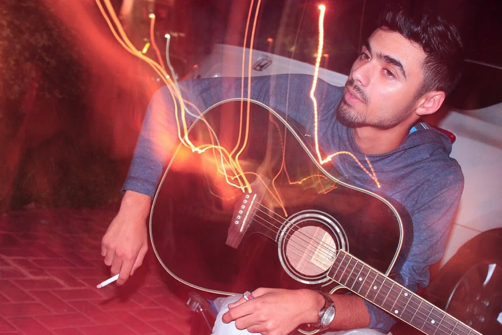 A man holding an acoustic guitar and a cigarette, with orange light trails.