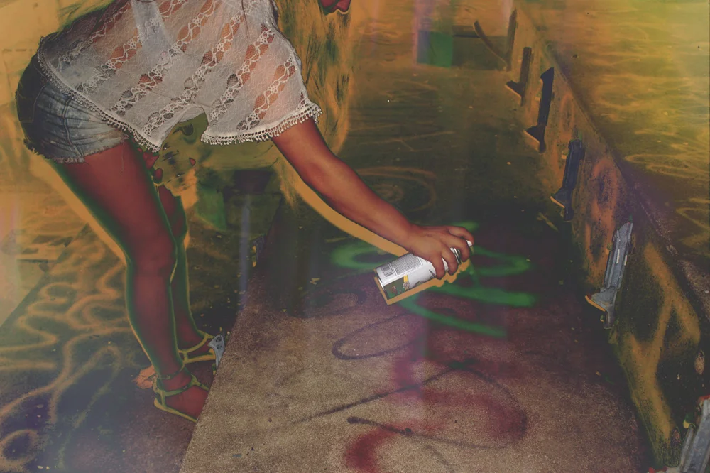 A person in a lace top and shorts spray painting the floor of a graffiti-covered room.