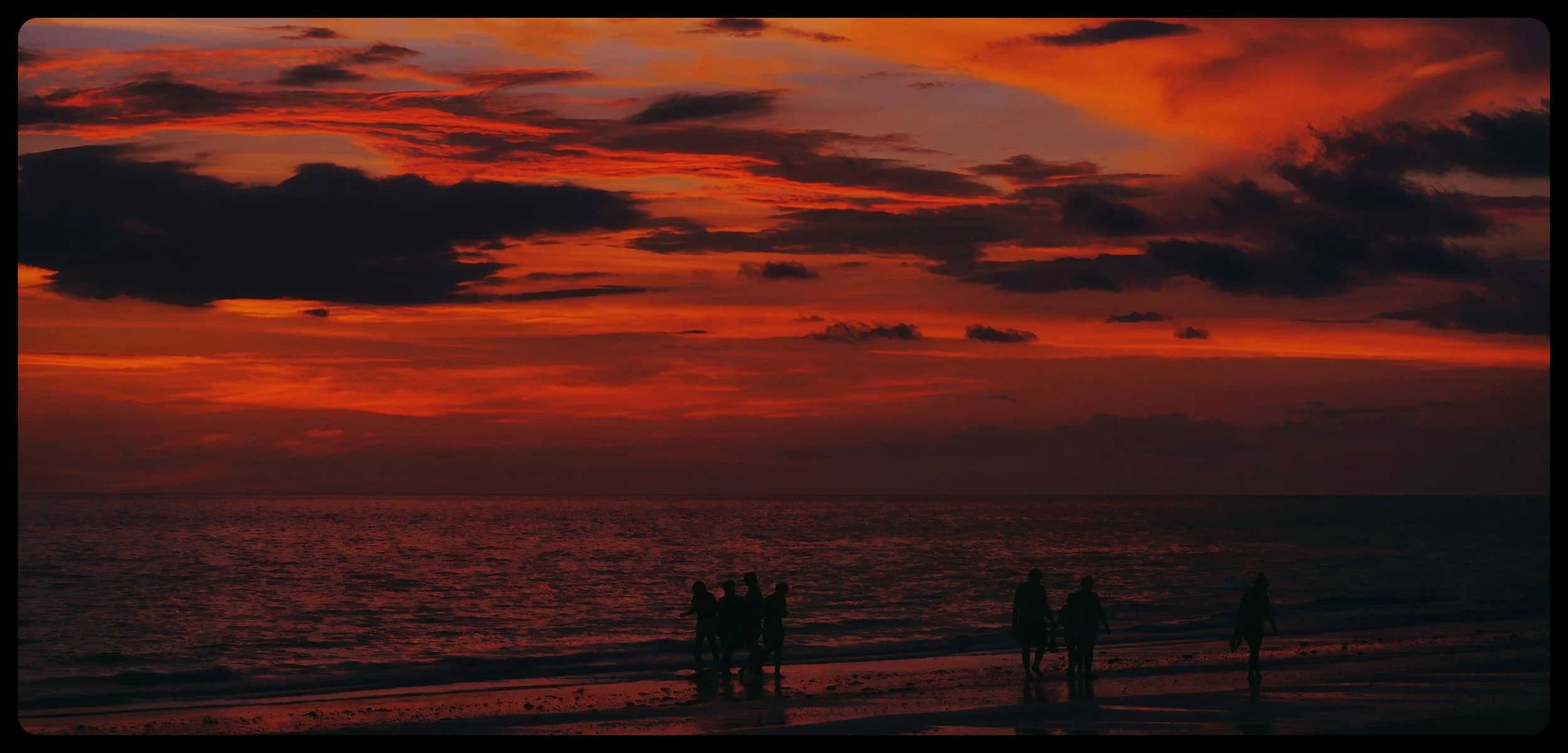 Silhouettes of people walking on a beach at sunset, with vibrant orange and red clouds in the sky and the ocean reflecting the colors.